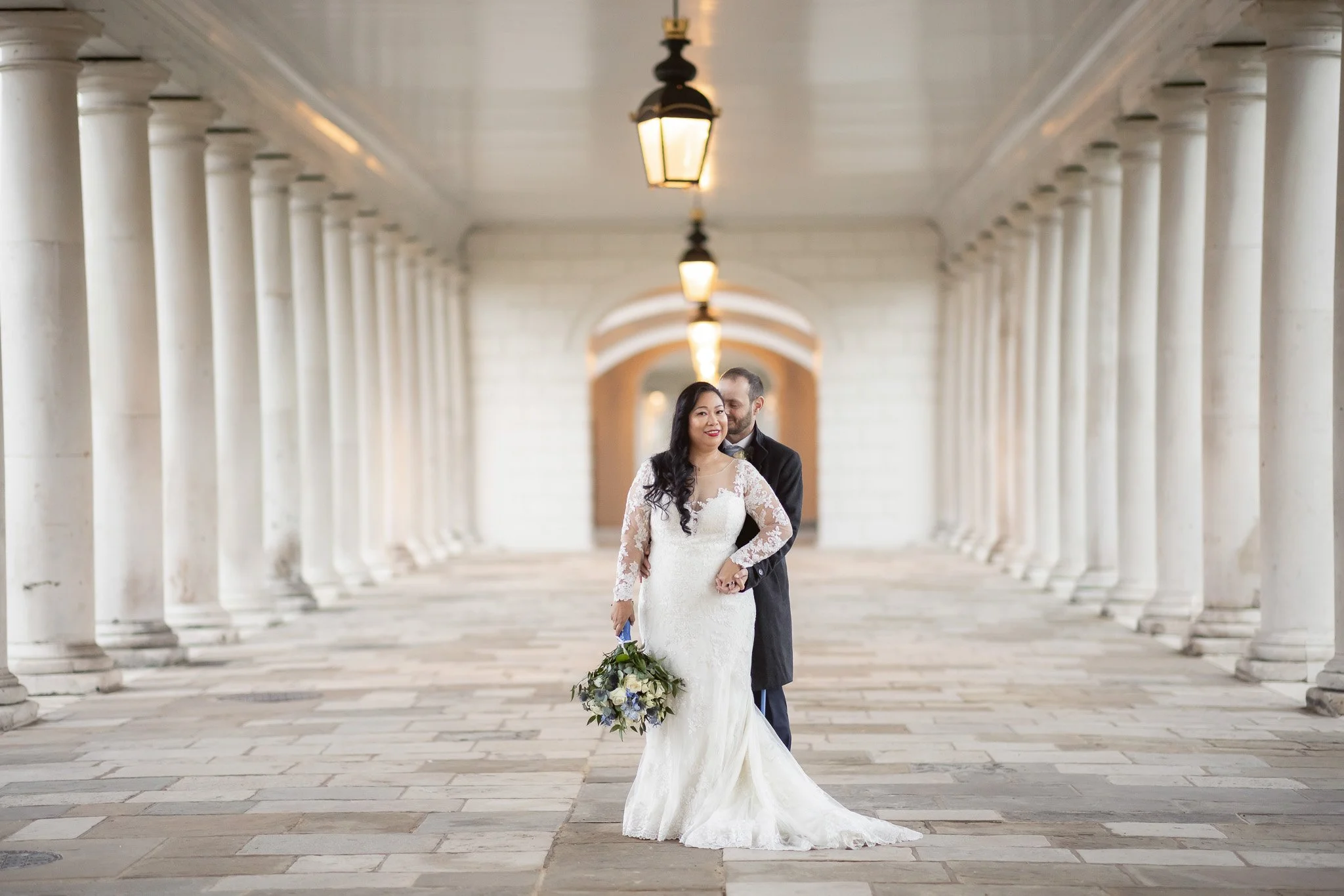 A bride in a white lace wedding dress holding a bouquet of flowers stands with a groom in a dark coat in a colonnaded walkway with hanging lamps.