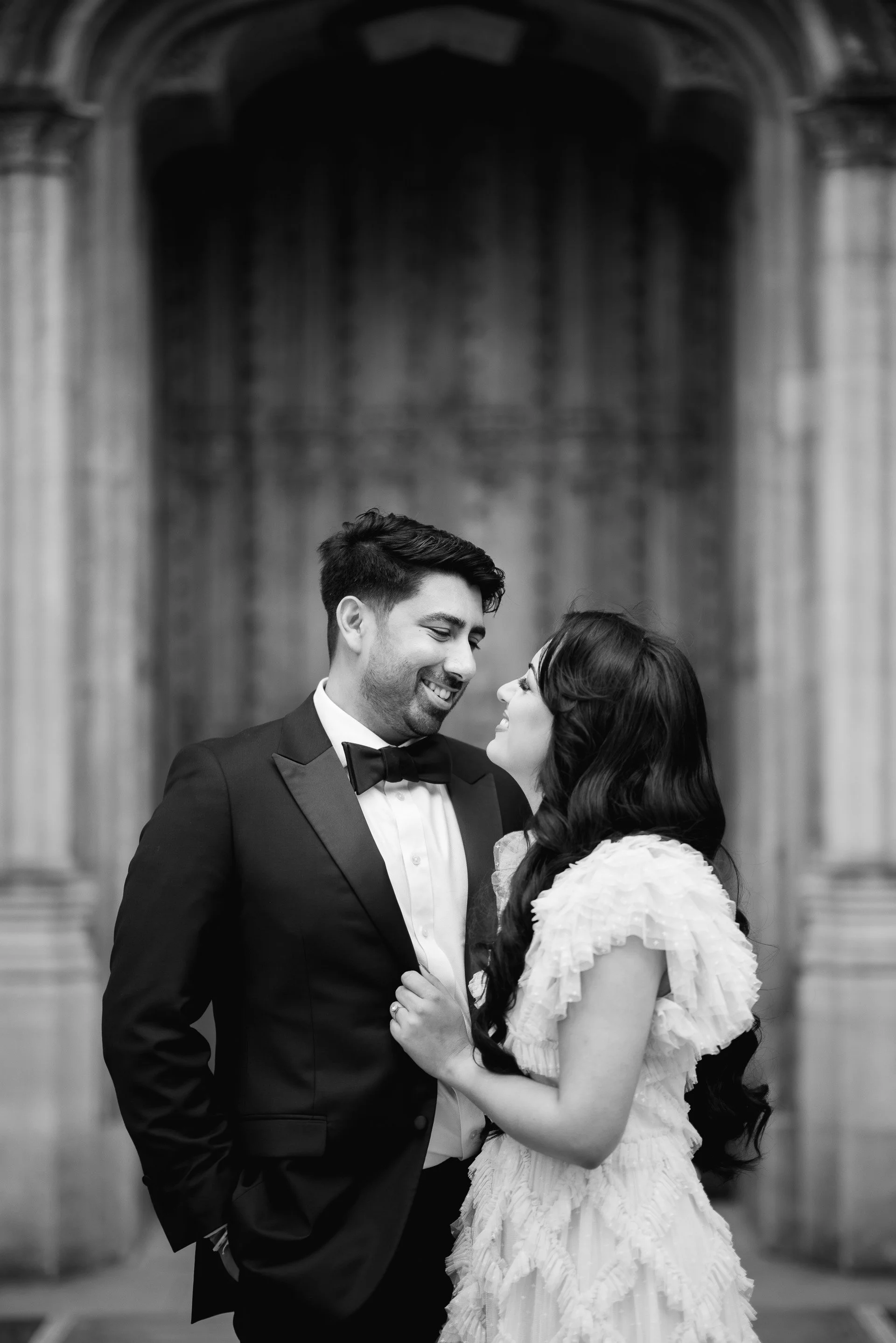 Black and white photo of a smiling man in a tuxedo and a woman in a white dress looking at each other in front of a large stone archway.