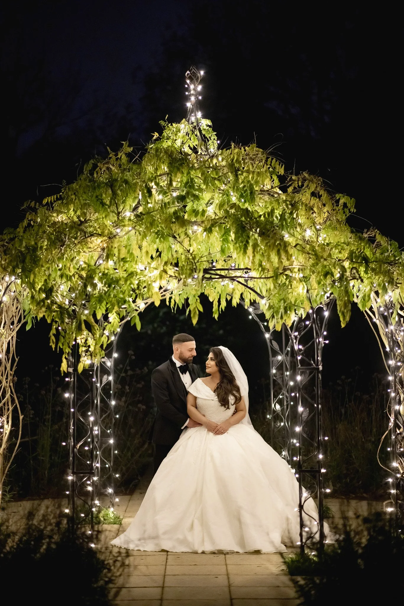 A bride and groom stand under an illuminated greenery archway at night, holding hands and looking into each other's eyes.