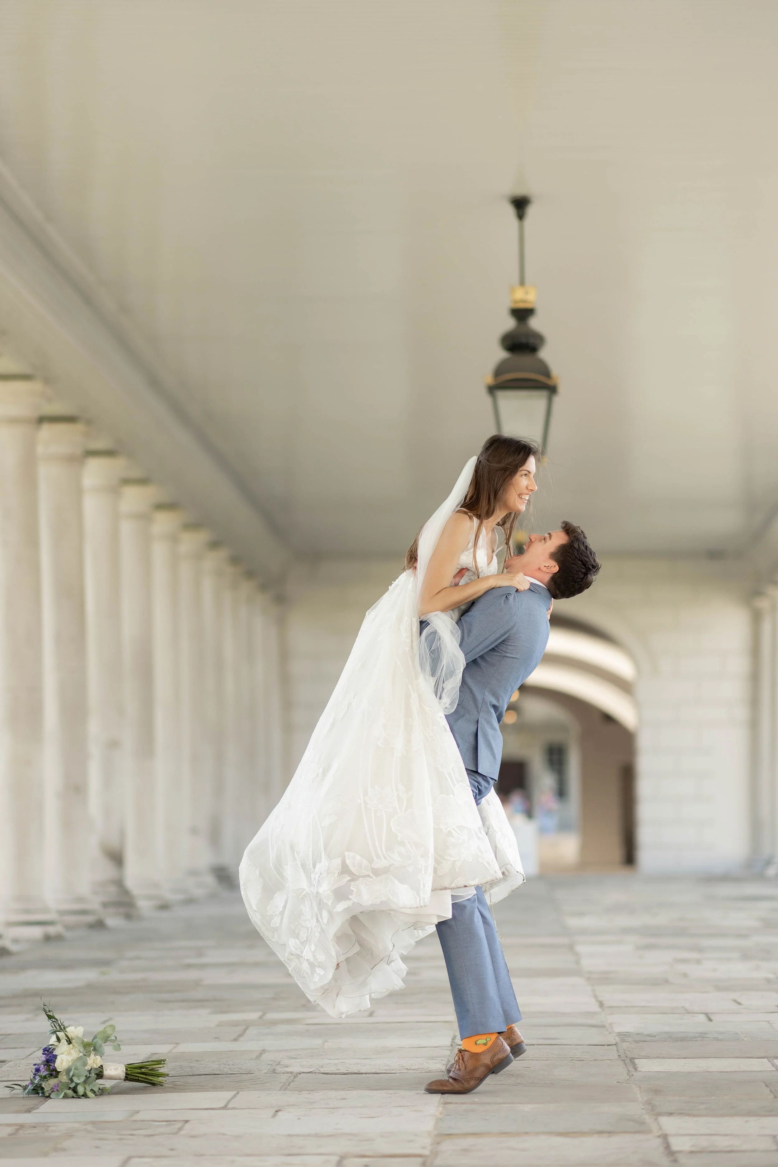 A couple in wedding attire, with the groom lifting the bride in an outdoor corridor with columns and lanterns, and a bouquet on the ground.