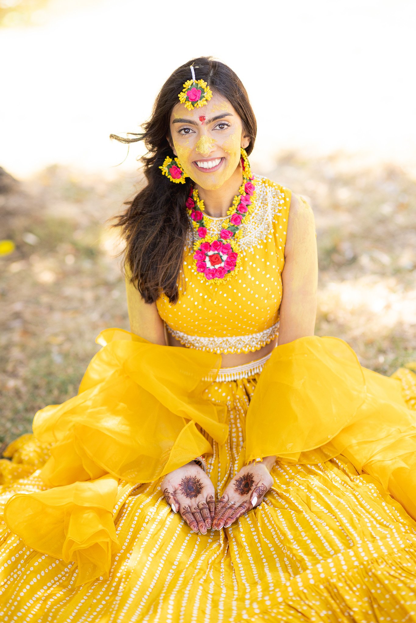 Woman dressed in a bright yellow traditional Indian outfit, sitting on the ground outdoors, smiling, with henna on her hands, and floral jewelry and decorations on her face.