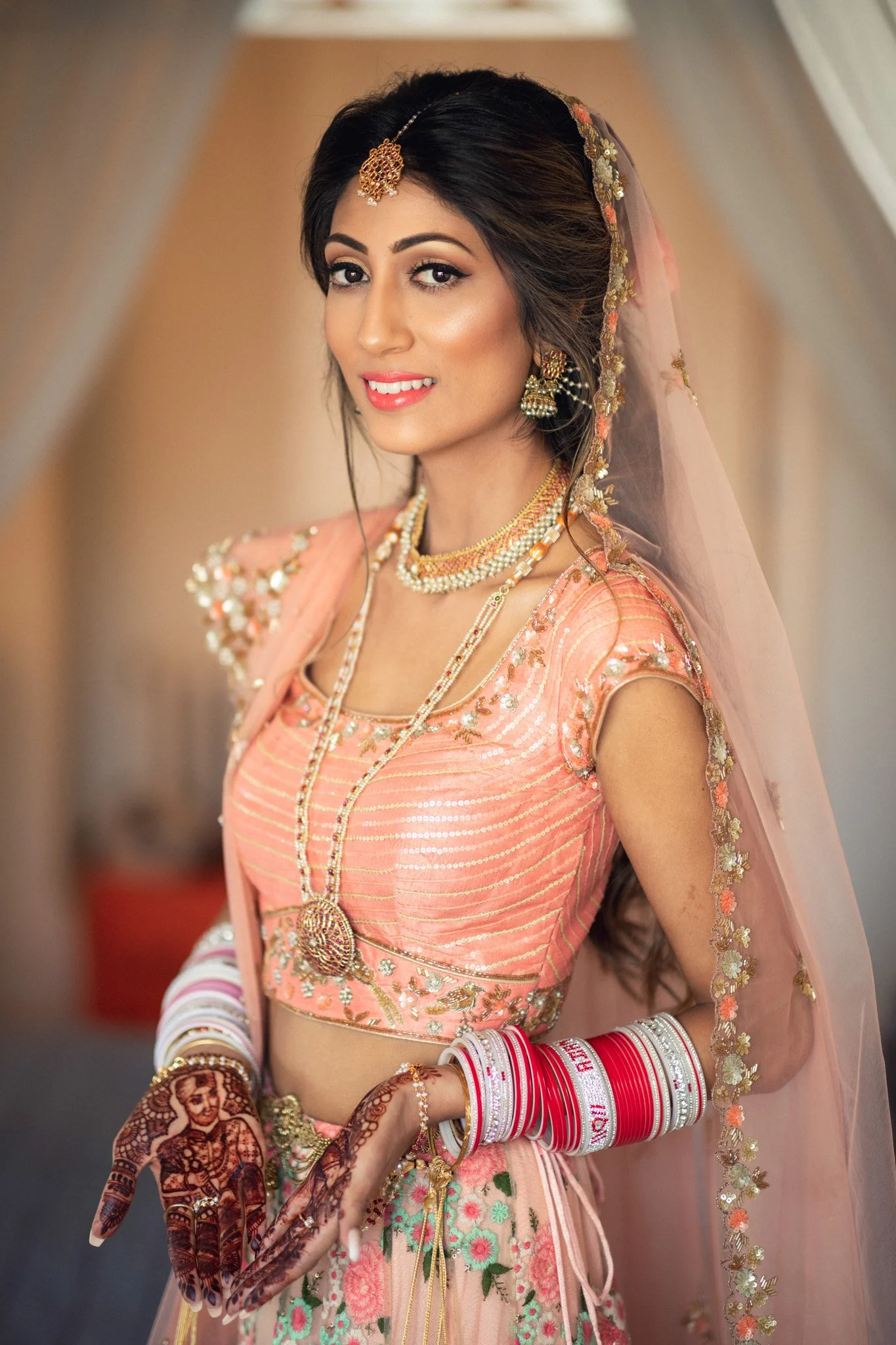 A woman in traditional Indian bridal attire with a peach and gold embroidered blouse and skirt, wearing jewelry including necklaces, earrings, and a maang tikka, with red and white bangles, henna on her hands, and a sheer peach dupatta with floral embroidery.