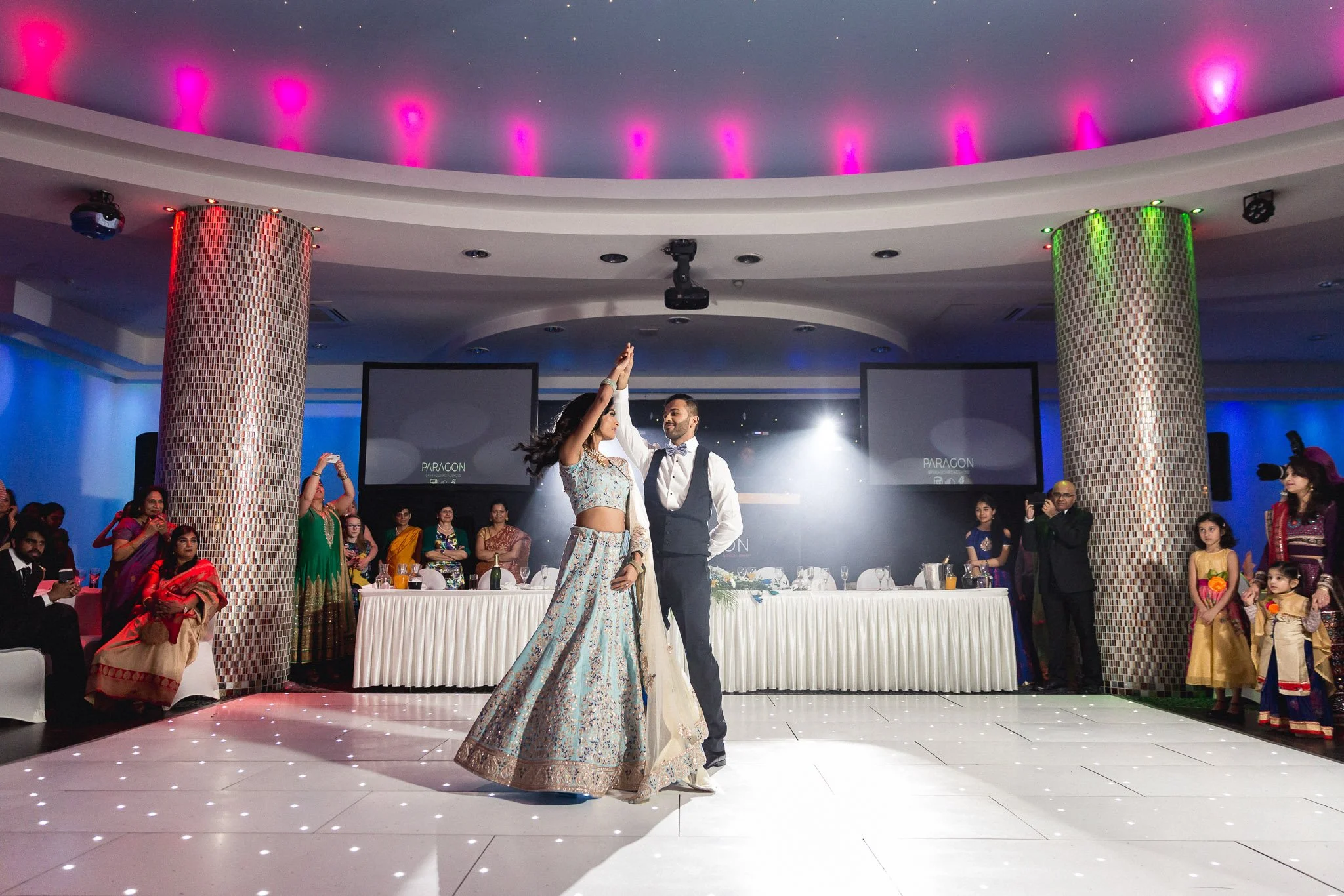 A couple dancing at a celebration with guests watching in a decorated hall with colorful lights and large screens.