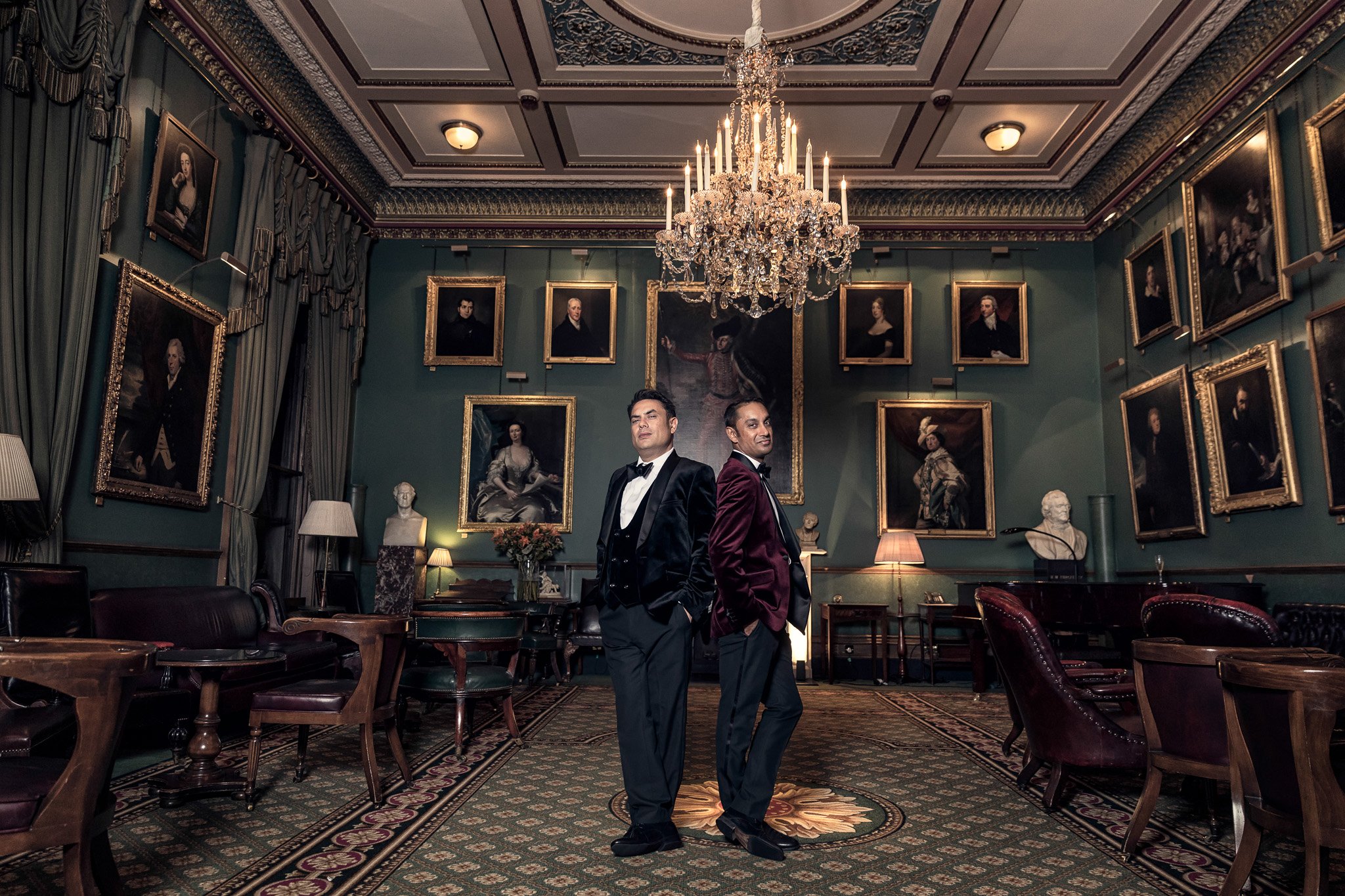 Two men in tuxedos posing in a luxurious, vintage-style room with framed portraits on teal walls, ornate chandelier, and antique furniture.