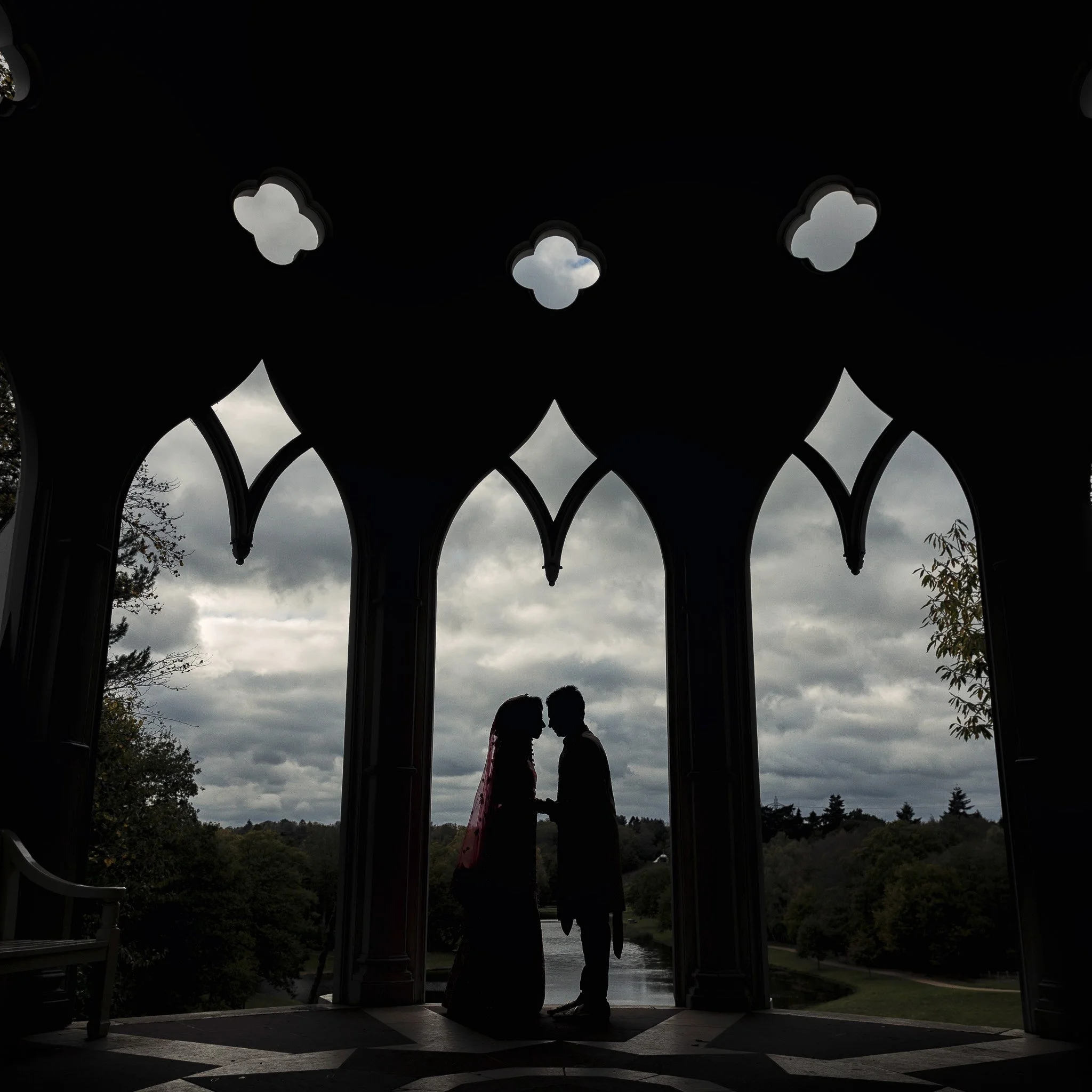 Silhouette of a couple holding hands and leaning close under an ornate pavilion with a cloudy sky and trees in the background.