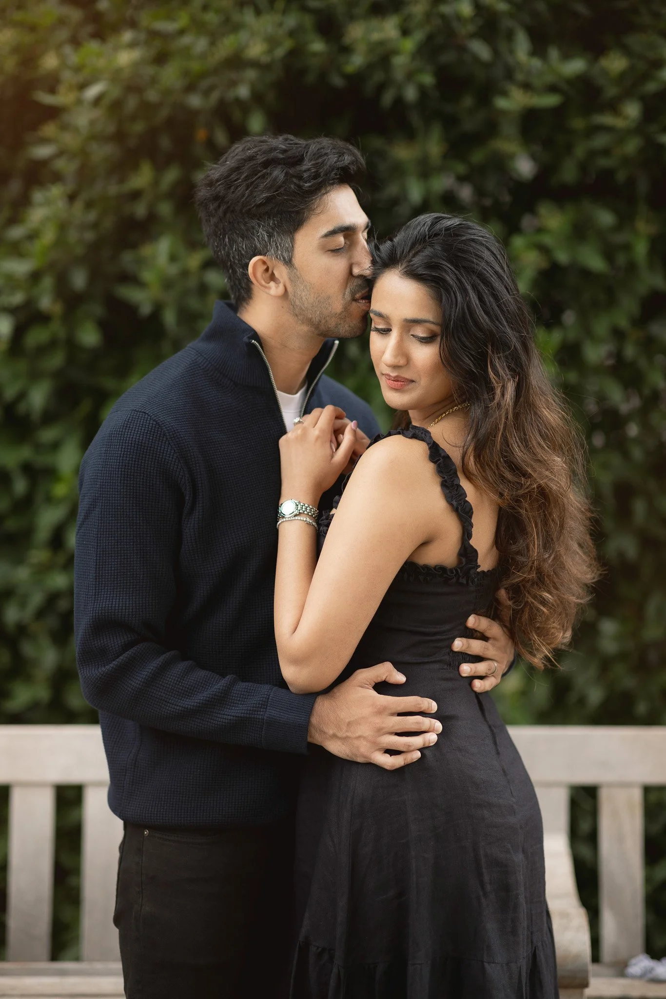 A young couple embracing outdoors, the man gently kissing the woman's forehead while she closes her eyes, with a garden and greenery in the background.