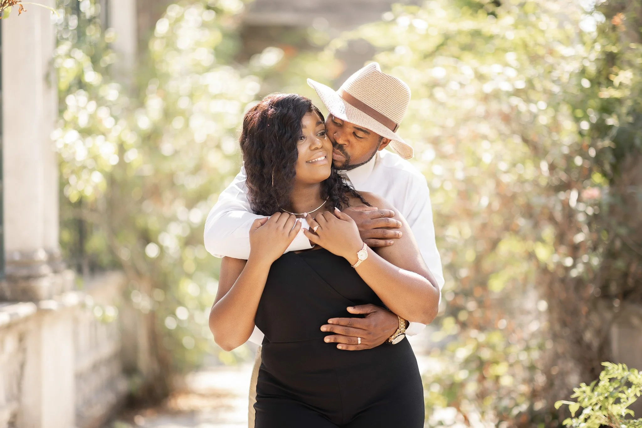 A man in a white shirt and a straw hat embracing a woman in a black dress outdoors, surrounded by greenery and sunlight.