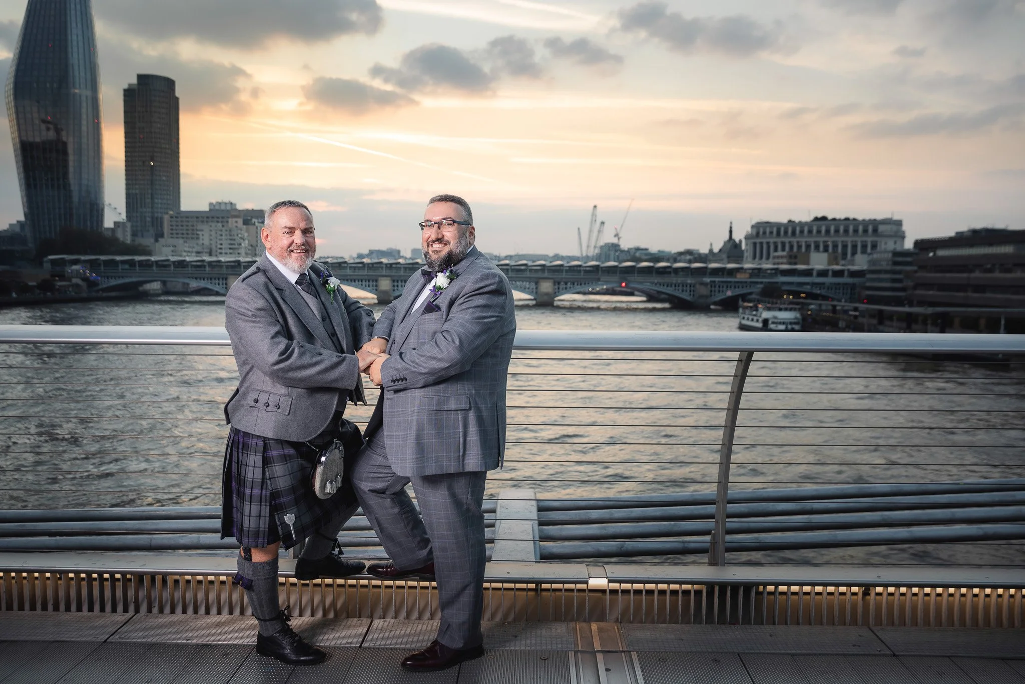 Two men in suits holding hands and smiling on a city bridge by the river during sunset, with modern buildings and a cloudy sky in the background.