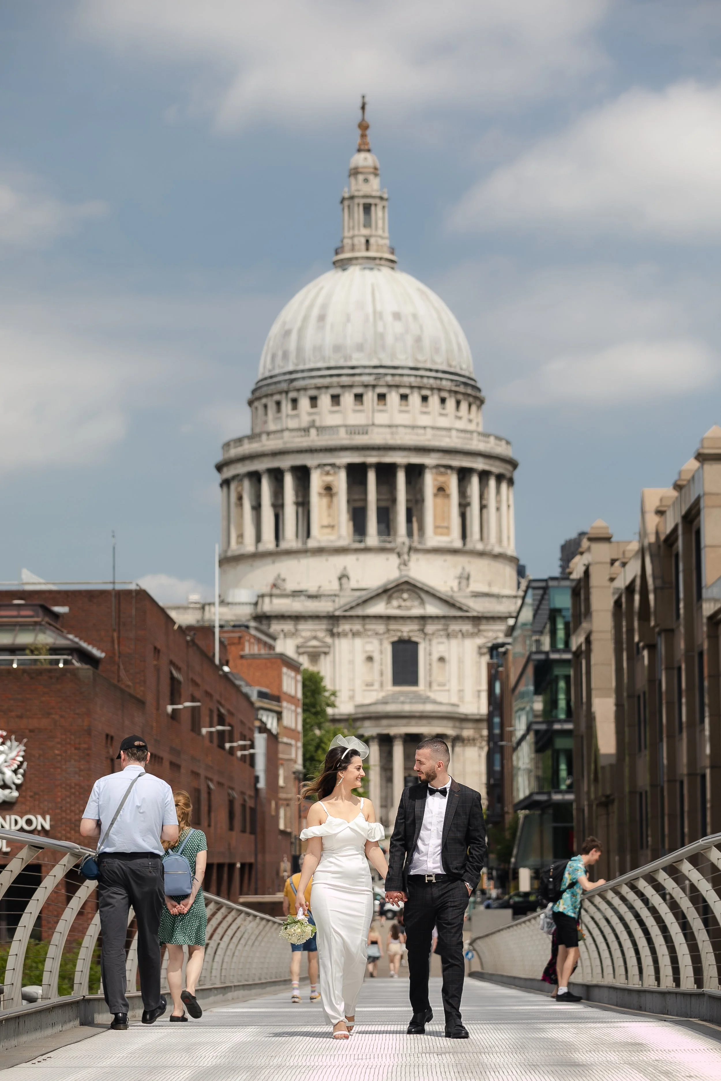 A newlywed couple walking hand in hand across a pedestrian bridge in London, with St. Paul's Cathedral in the background. The bride is wearing a white wedding dress, and the groom is dressed in a dark suit with a bowtie.