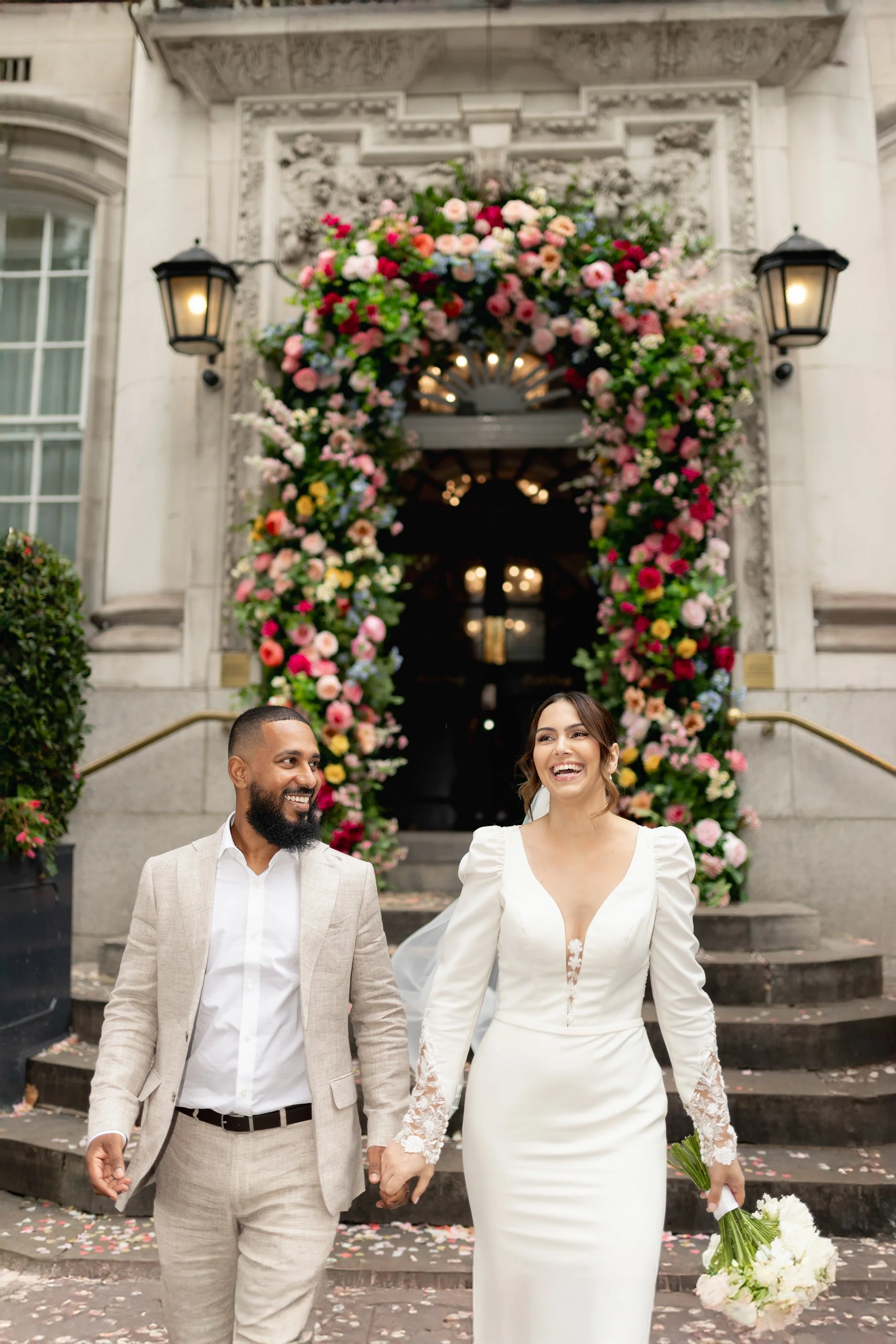 A newly married couple, a man and woman, walking hand in hand out of a building decorated with a large arch of colorful flowers, smiling and happy, during their wedding celebration.