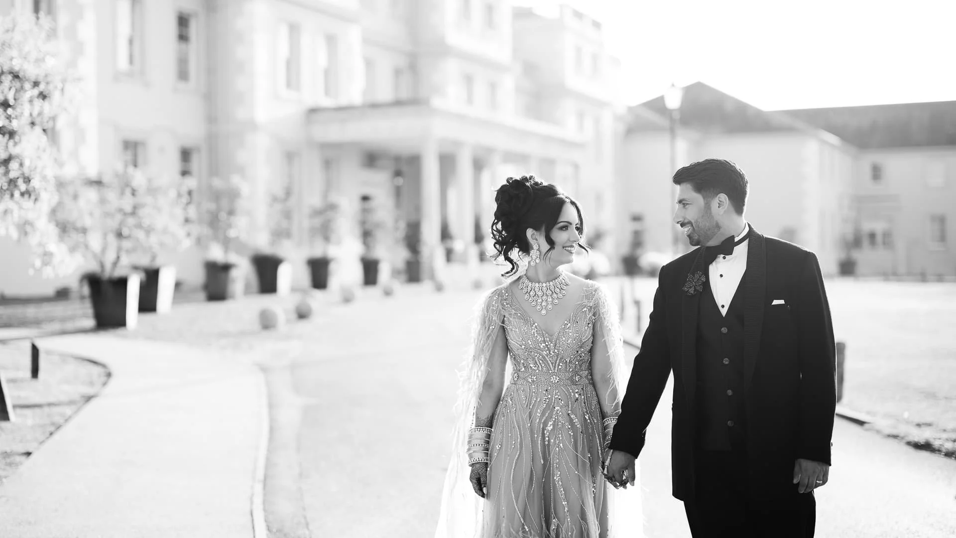 Black and white photo of a wedding couple holding hands and smiling at each other outside near buildings