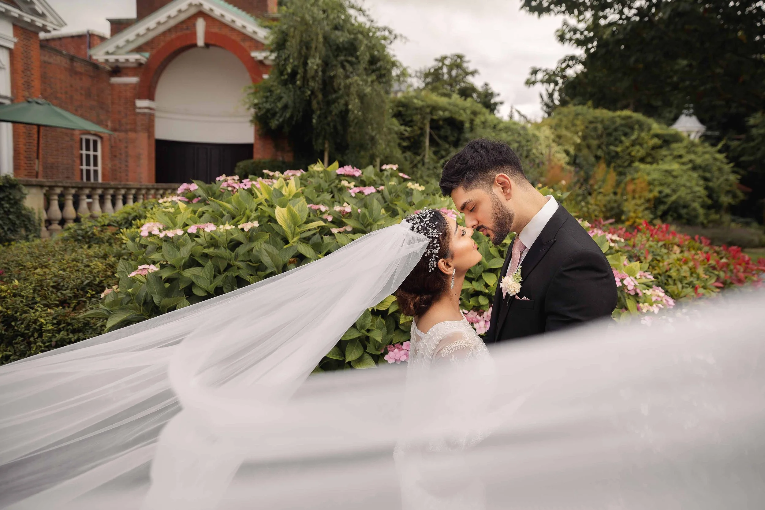 A bride and groom standing close, touching foreheads, surrounded by greenery and flowers, with a brick building in the background.