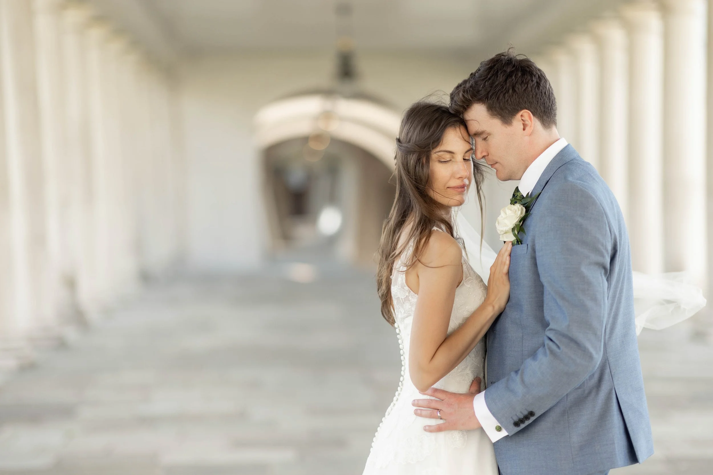 Bride and groom embrace in a tunnel with white columns, touching foreheads with eyes closed, romantic and tender moment.