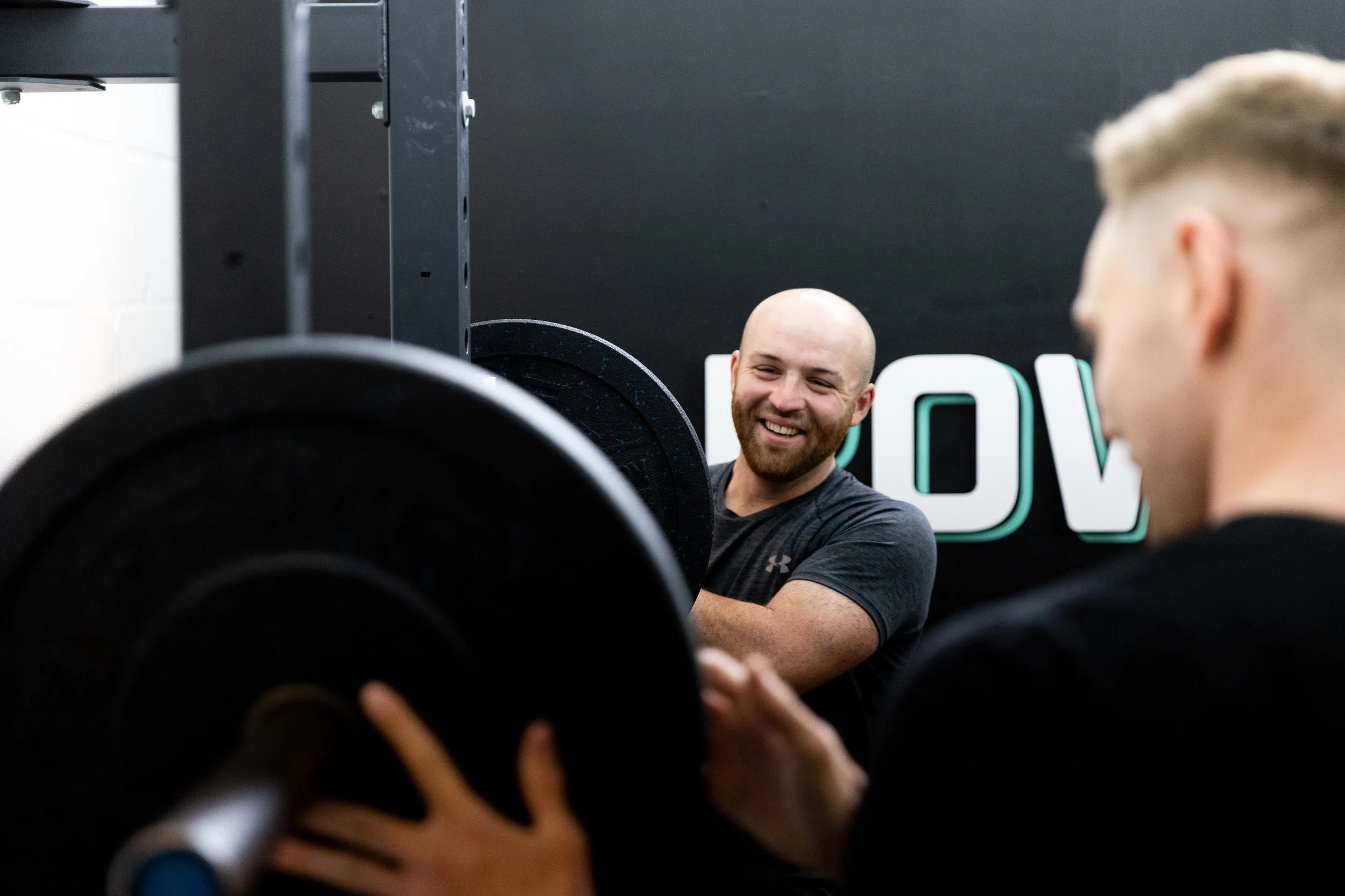 A man with a bald head and beard smiling as he observes a person with blond hair, who is partially out of frame, in a gym or fitness setting with weightlifting equipment.