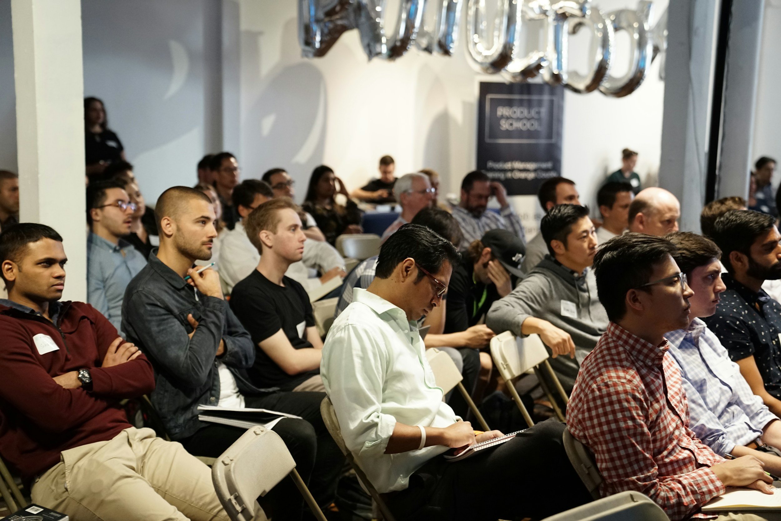 Audience attending a conference or seminar, seated attentively in a dimly lit room, with some taking notes and others listening, featuring a diverse group of men and women.