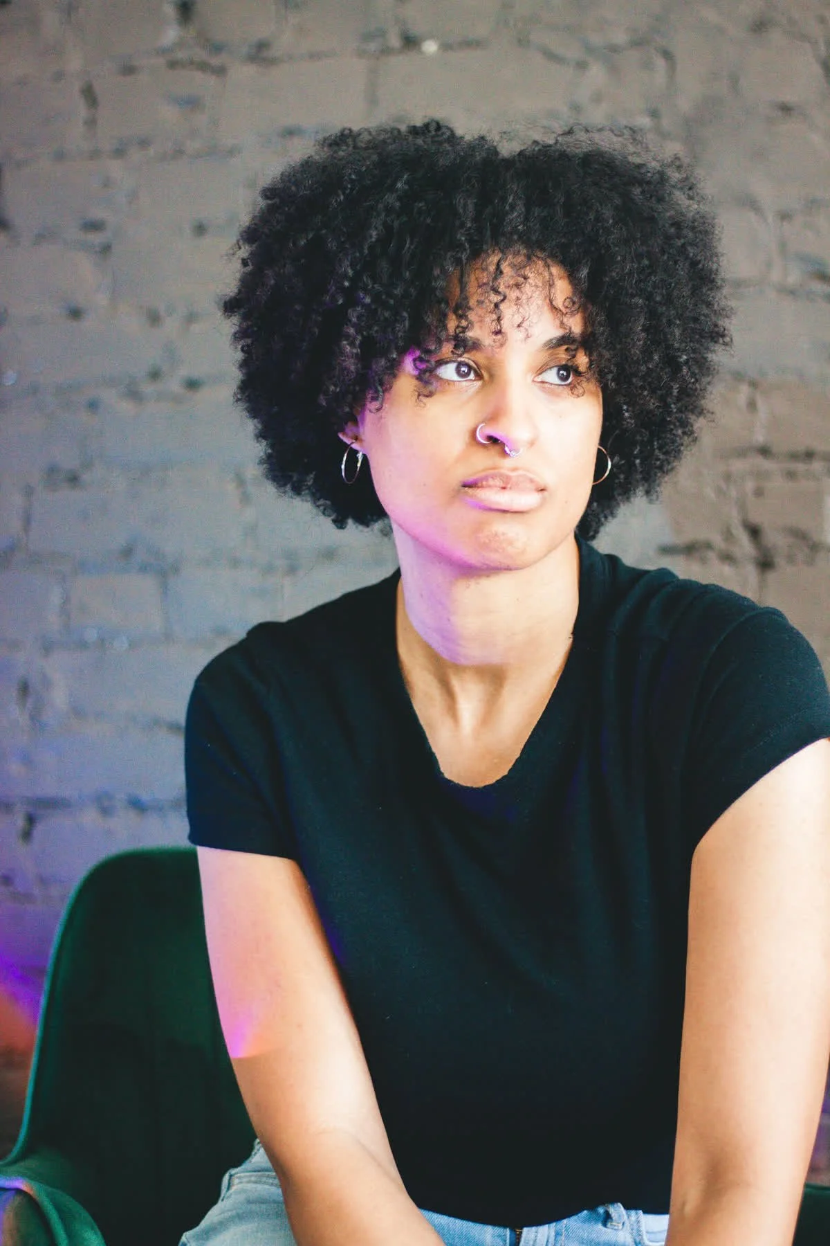 A young woman with black, curly hair, wearing hoop earrings and a nose ring, sitting against a gray brick wall, looking to the side.