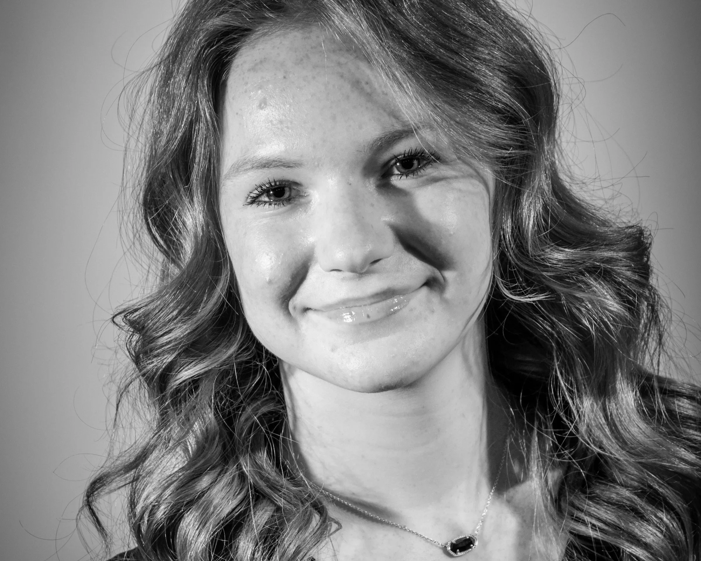 A black and white portrait of a young woman with wavy hair smiling slightly, wearing a necklace with a dark stone pendant.