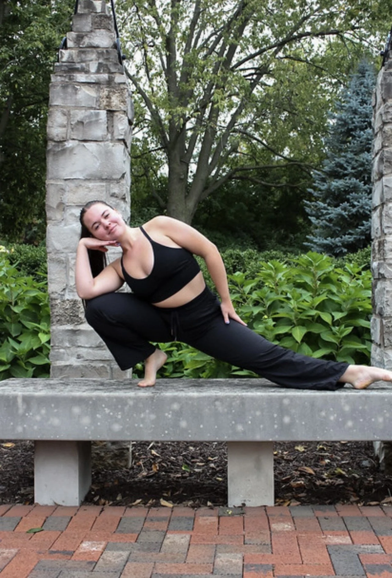 A woman practicing yoga outdoors on a stone bench, doing a side lunge pose with a relaxed expression, surrounded by green foliage and trees.