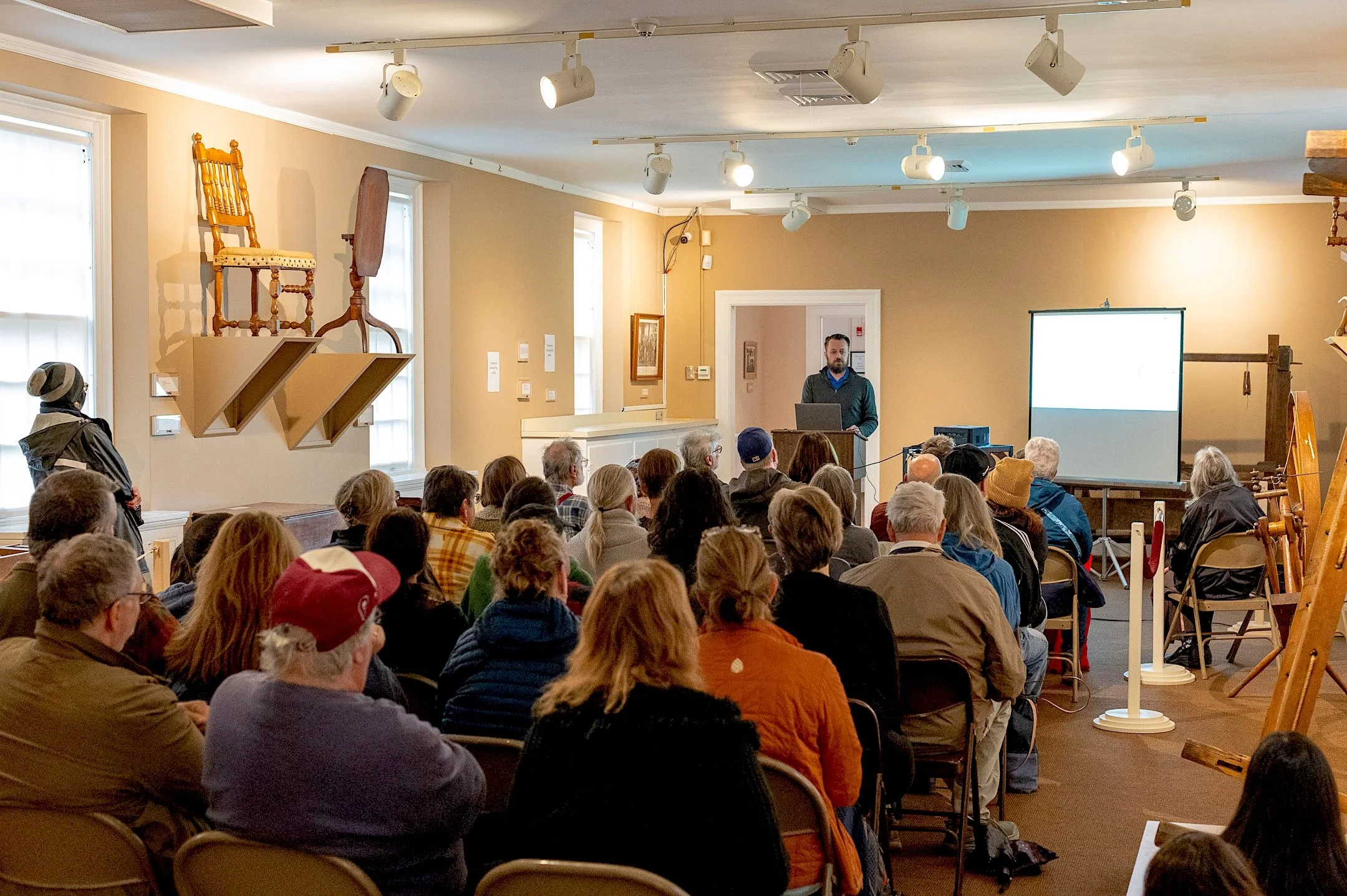 A man giving a presentation at an indoor event to an audience seated in a room with artwork on the walls and antique furniture displayed on shelves.