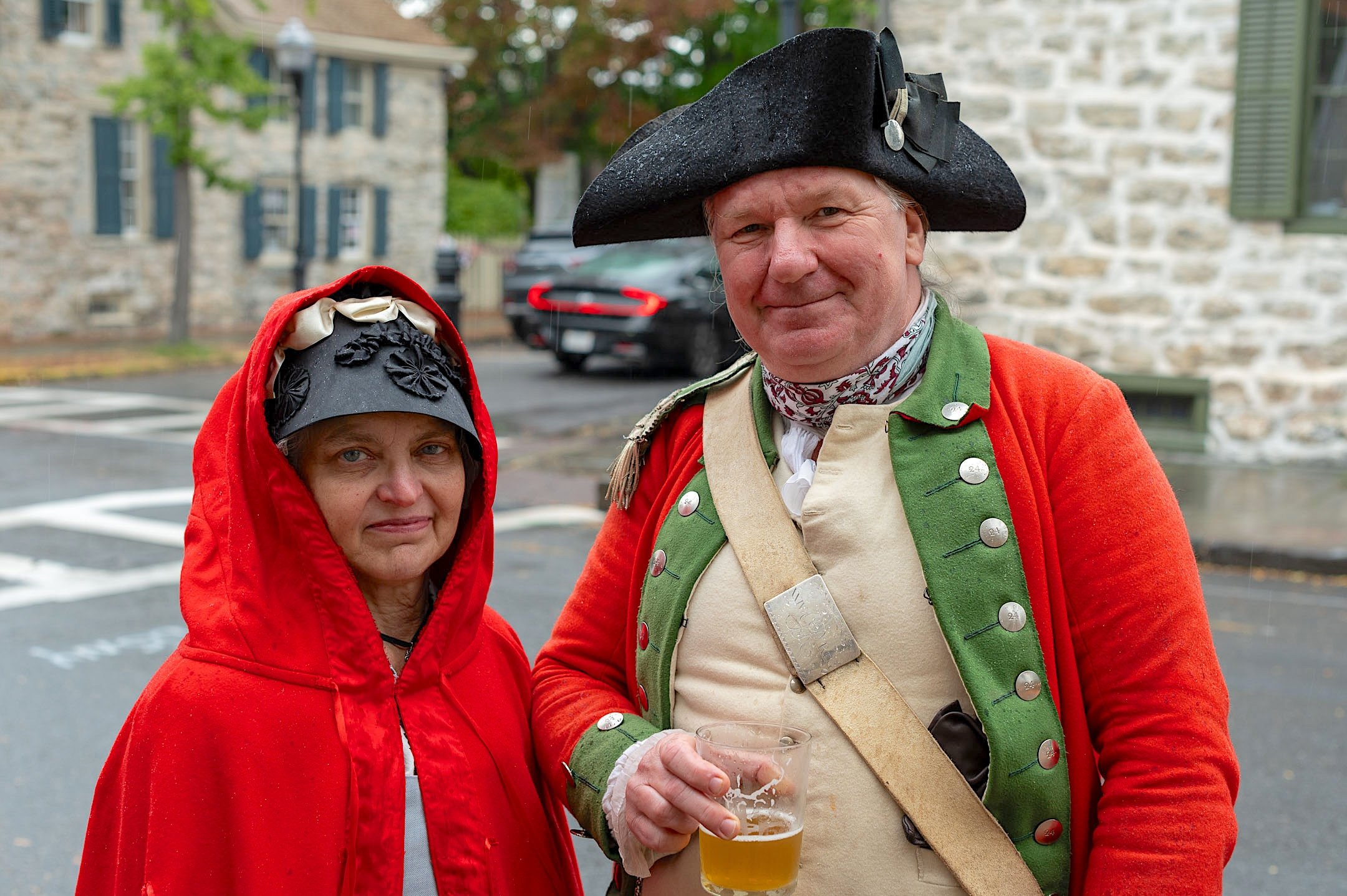 A woman in a red hooded cloak and a man in a historical military uniform holding a glass of beer standing outdoors in a parking lot with stone buildings in the background.