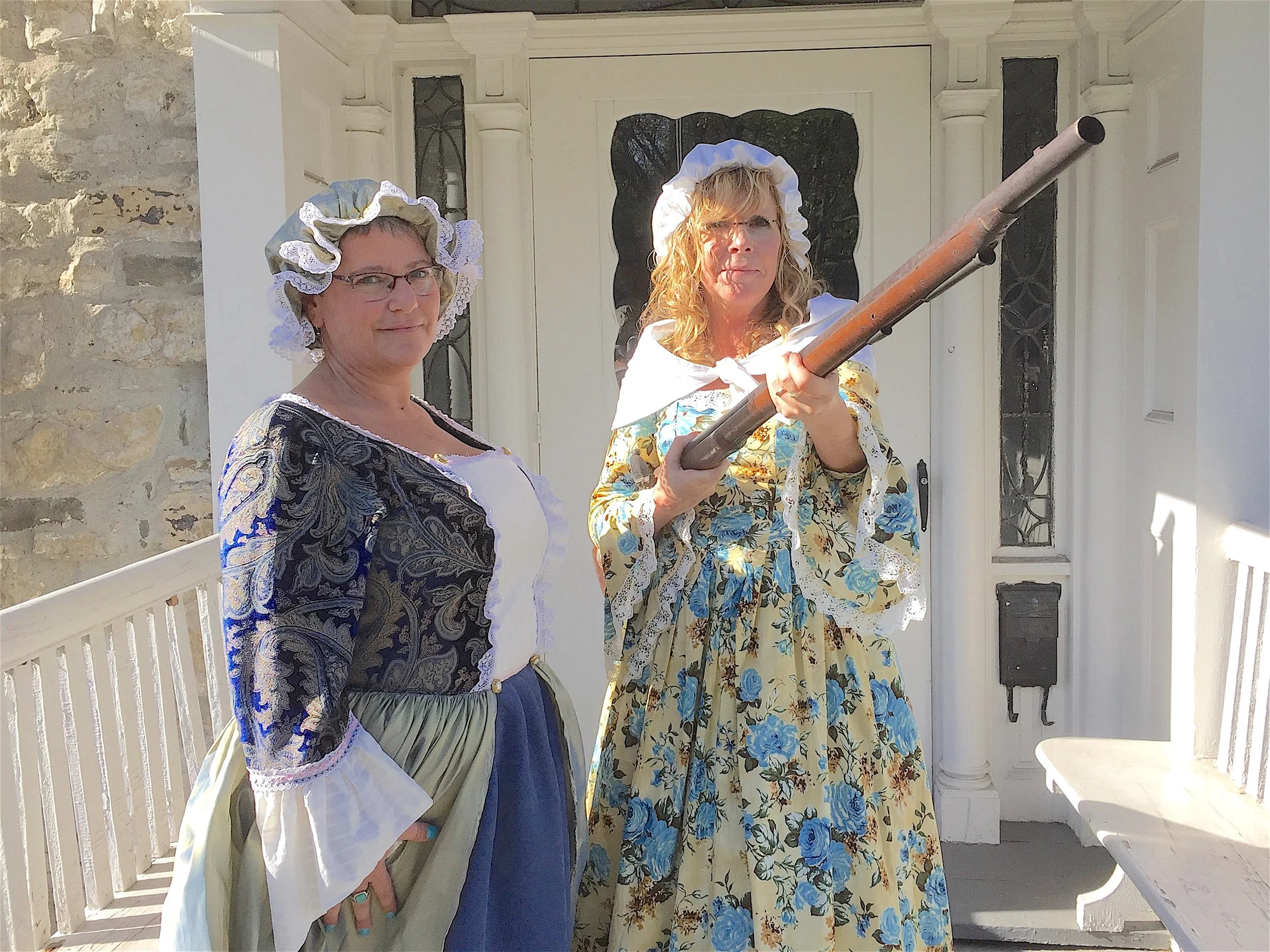 Two women dressed in historical costumes, one holding a musket and the other standing beside her, outside a house with a white door and stone wall, during daytime.