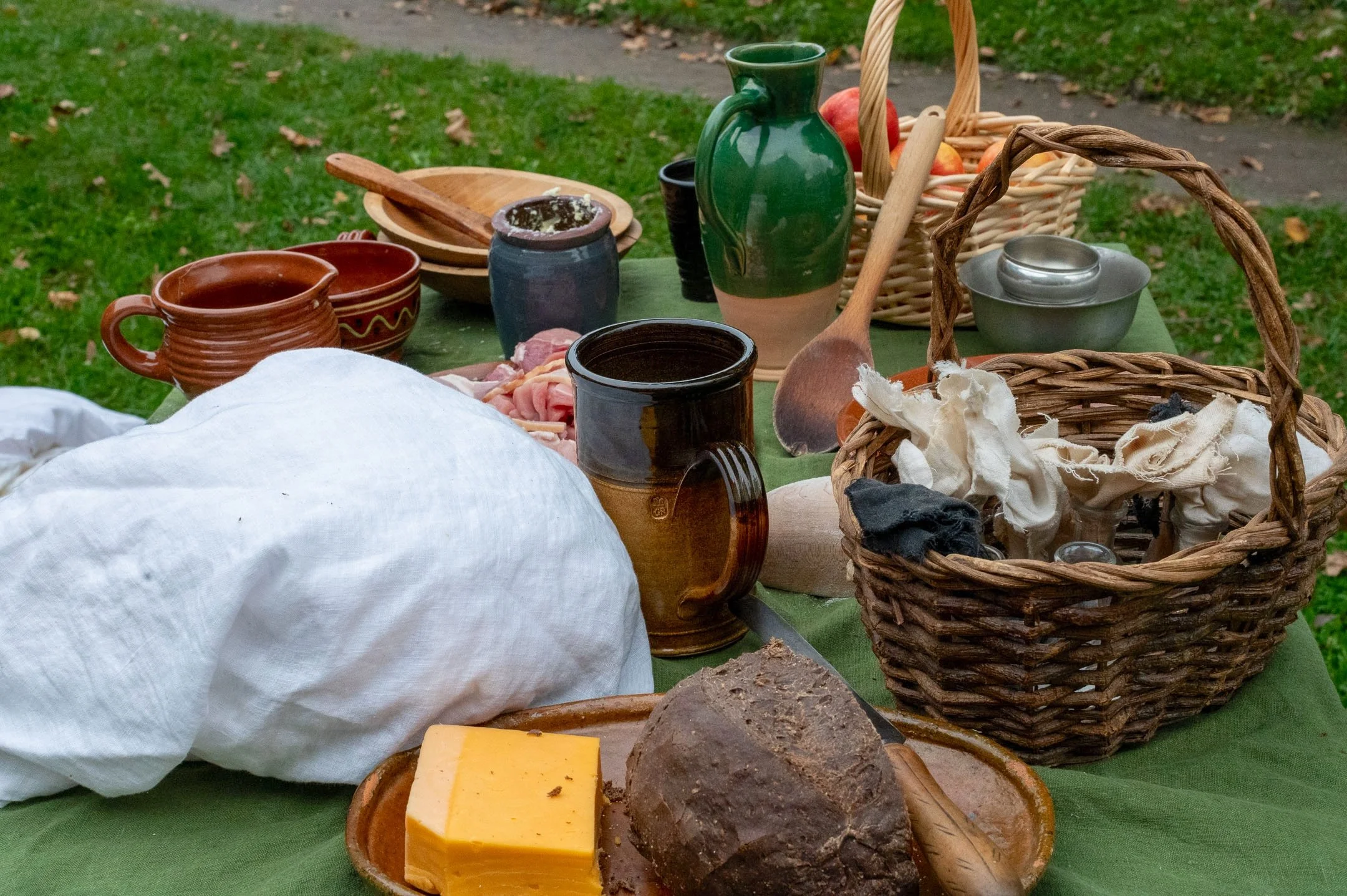 A rustic outdoor table set with cheese, bread, pottery, a basket of cloth, and various dishware on a green tablecloth, with grass and a walkway in the background.