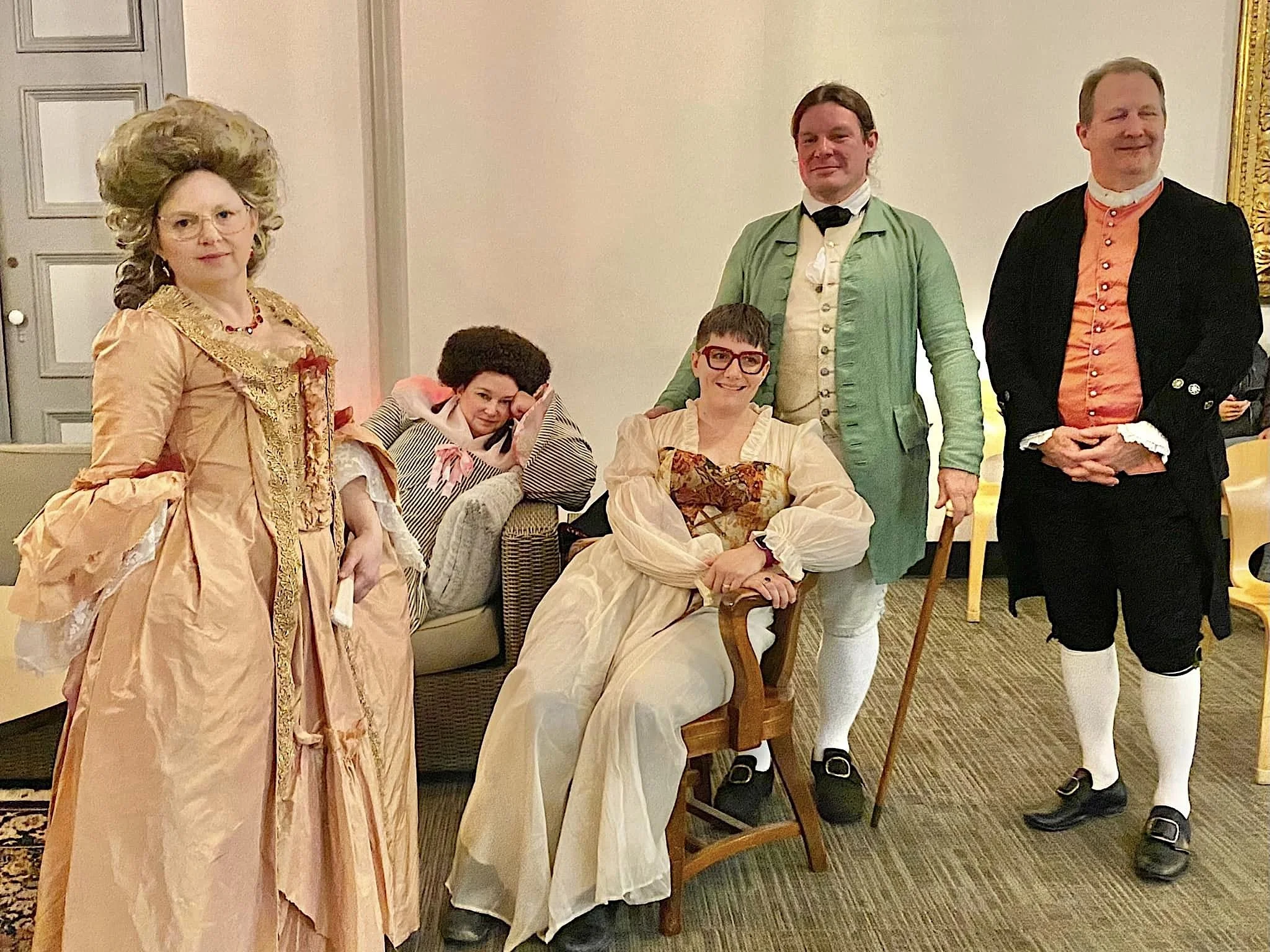 Group of six people dressed in 18th Century historical costumes, posing indoors in a room with beige walls and a carpeted floor.