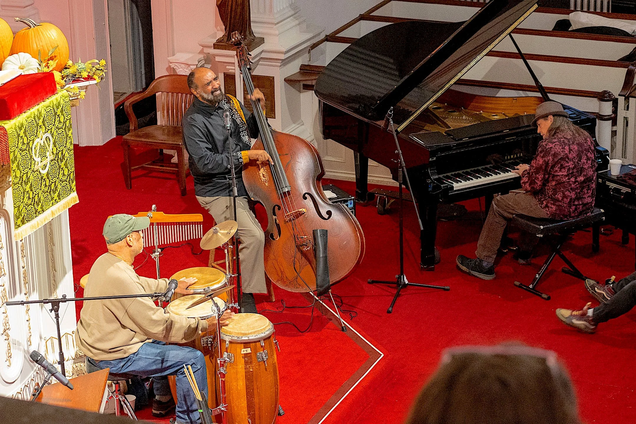 Jazz band performing on stage with a pianist, double bassist, and drummer in a room with red carpet, white walls, and decorative elements.