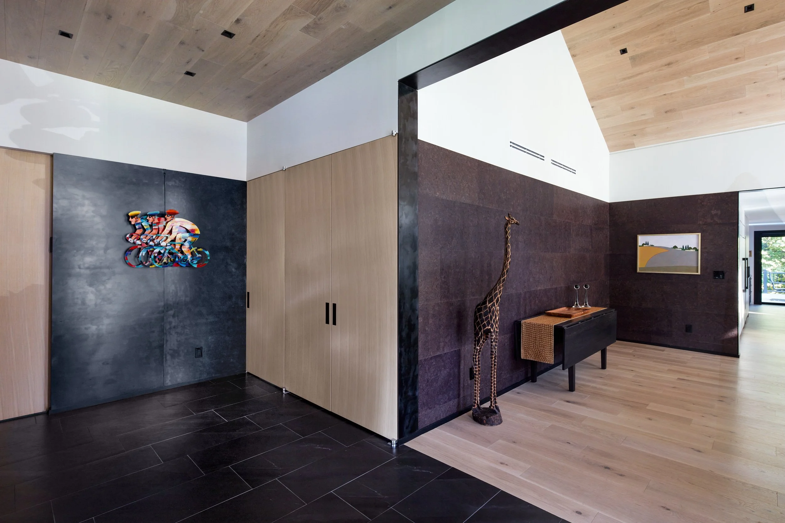 Interior foyer of new house renovation. Dark hardwood floors on the left, light hardwood on the right leading to the front door. Pale hardwood ceiling. View looks at a corner with artwork to either side.