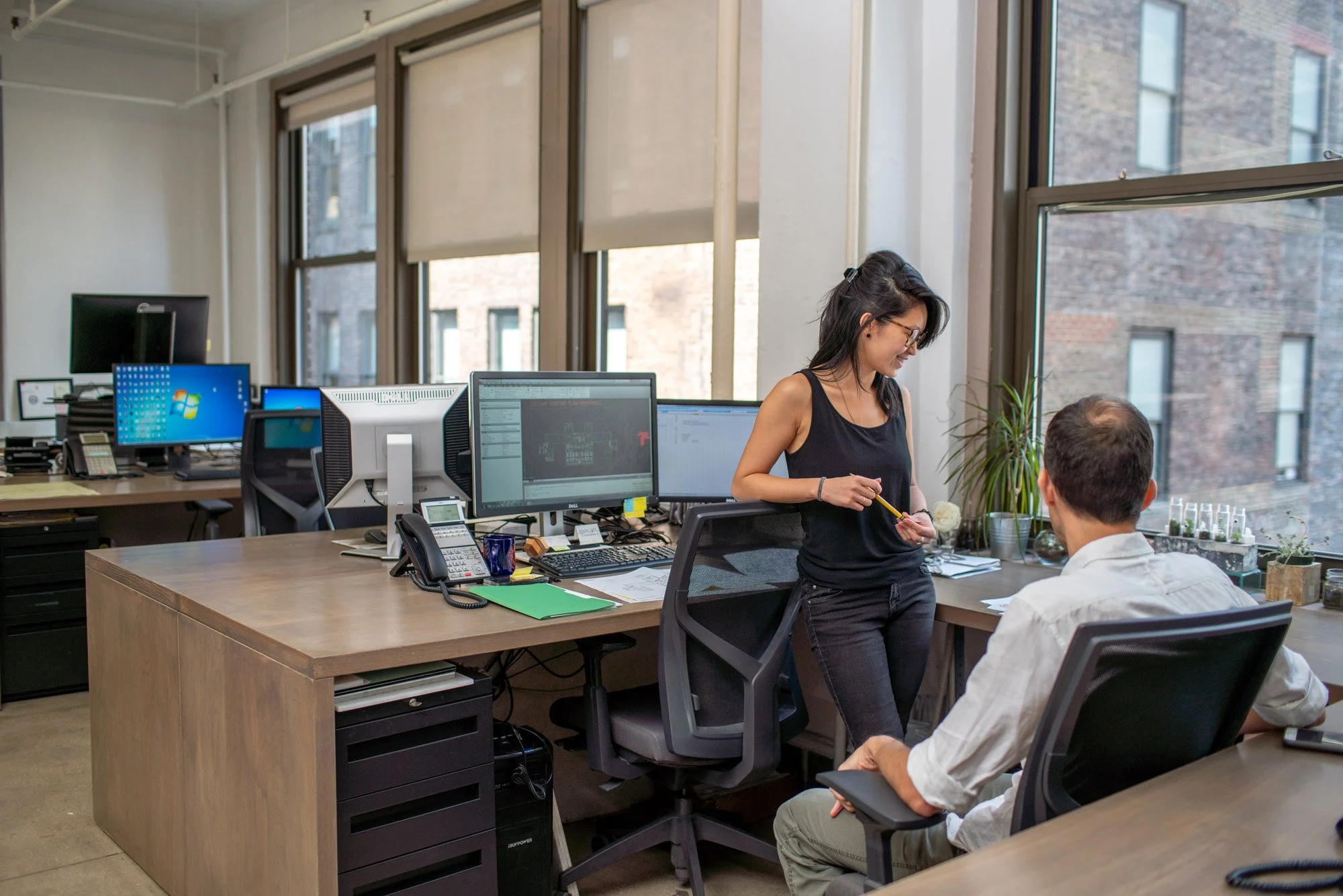 Two people having a conversation at a desk in a modern office with multiple computer monitors, office chairs, and windows with blinds.