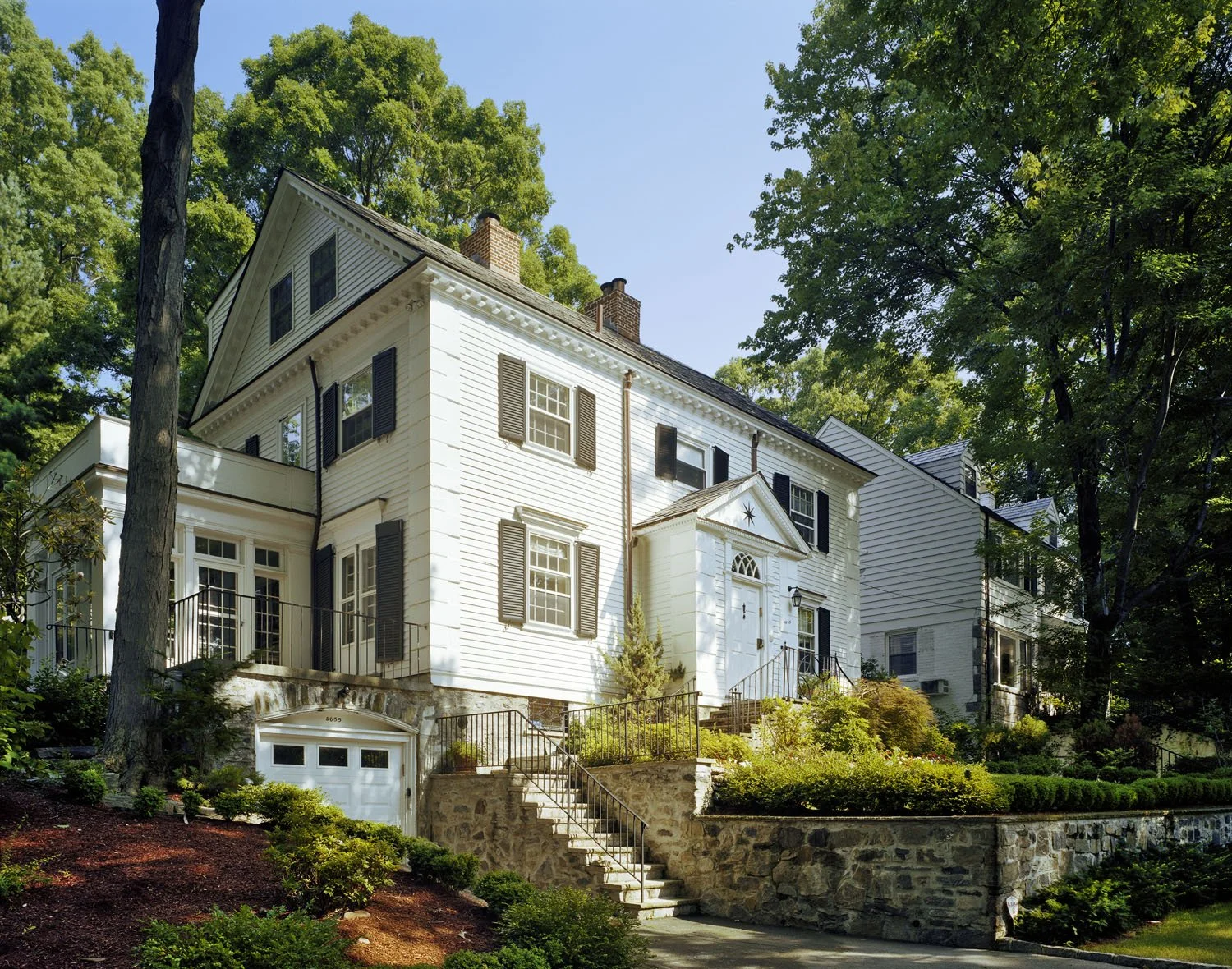 This exterior two story mansion sits atop a raised lawn and has lush foliage that brightens the white paneling.