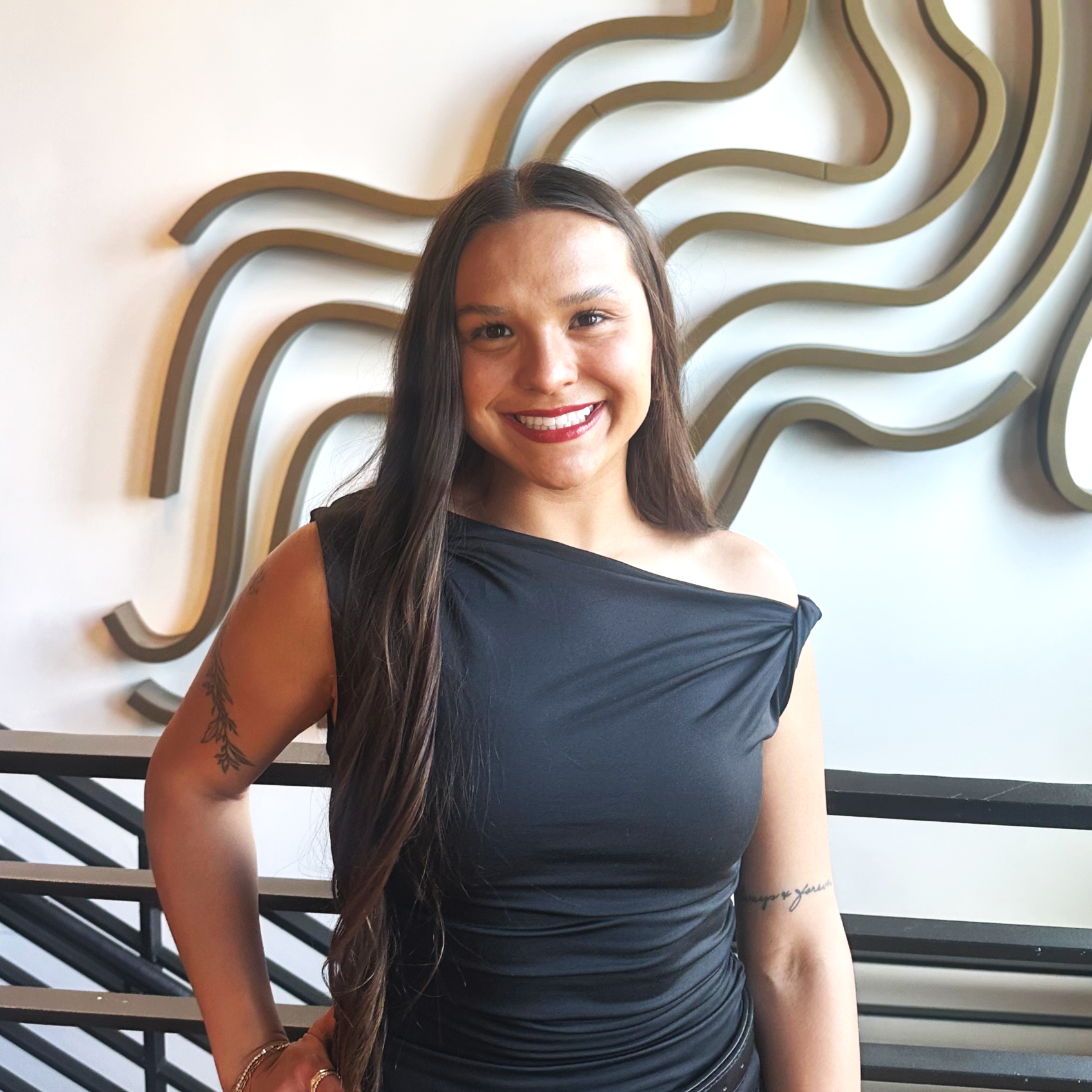 A woman with long brown hair, wearing a black ribbed turtleneck sweater and a gold necklace, smiling in front of a modern abstract wall decor.
