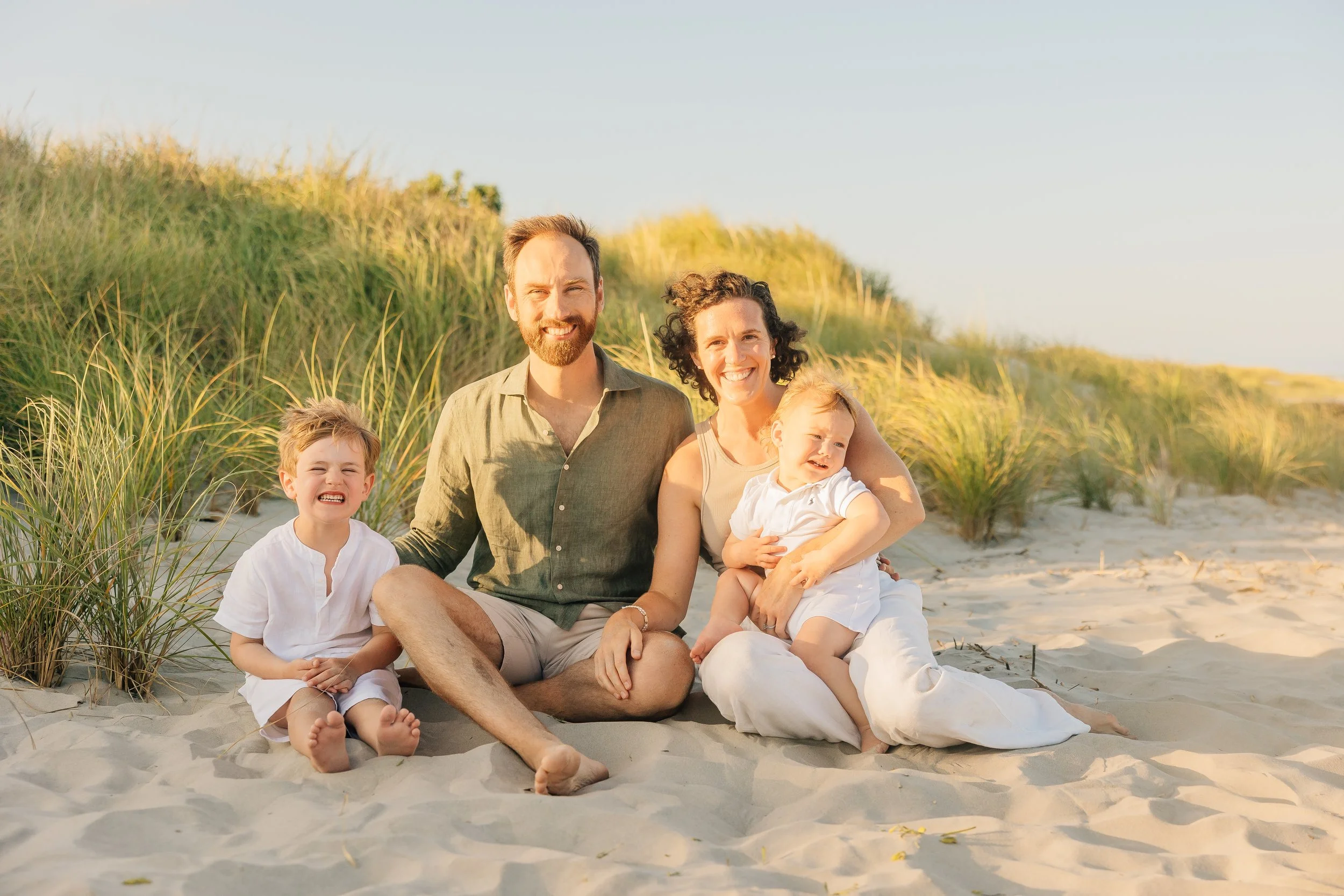 A happy family of four sitting on a sandy beach with grass and dunes in the background during sunset. The father, mother, and two young children are smiling and enjoying the moment.
