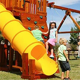 Children playing on a wooden playground set with a yellow tube slide and climbing wall.