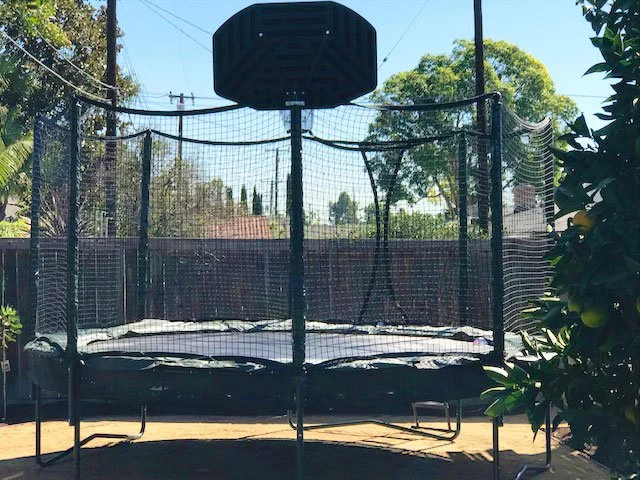 Outdoor trampoline with safety net and basketball hoop in a backyard setting.