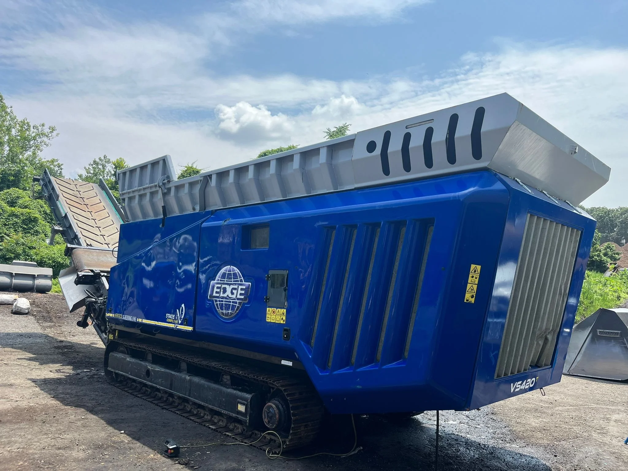 A large blue excavation or construction machine with tracks, labeled EDGE, is outdoors under a partly cloudy sky, with a piece of equipment or attachment on top.