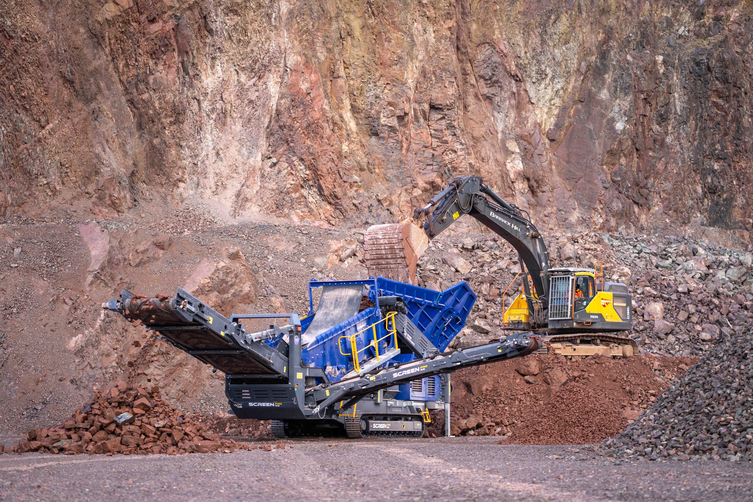 A large construction excavator loading rocks into a mobile rock crusher in a quarry.