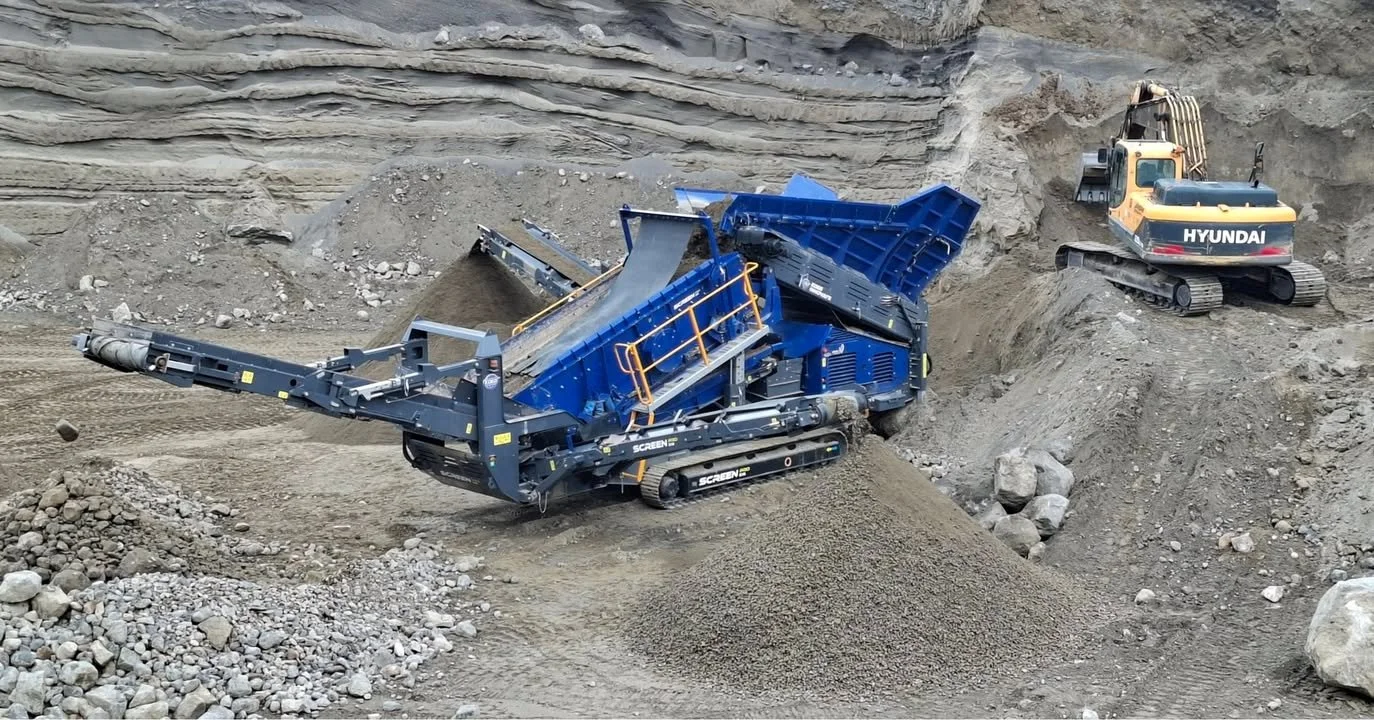 A large blue industrial rock crusher on tracks crushing rocks and gravel in a quarry with a yellow excavator in the background.