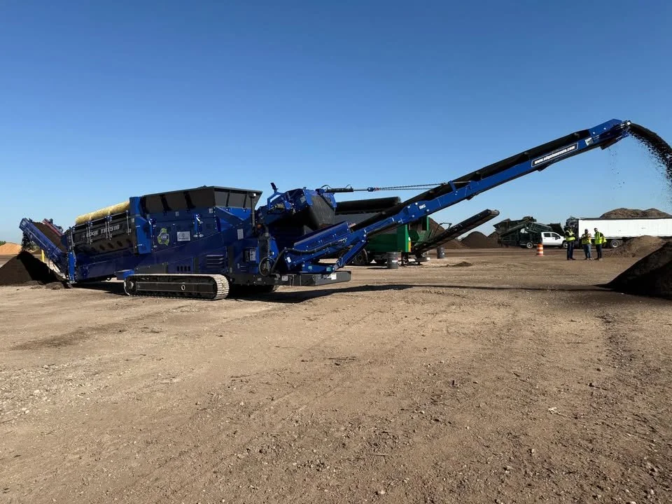 A large blue industrial machine operating on a dirt site, unloading or processing rocks or gravel, with a conveyor belt extending outward.