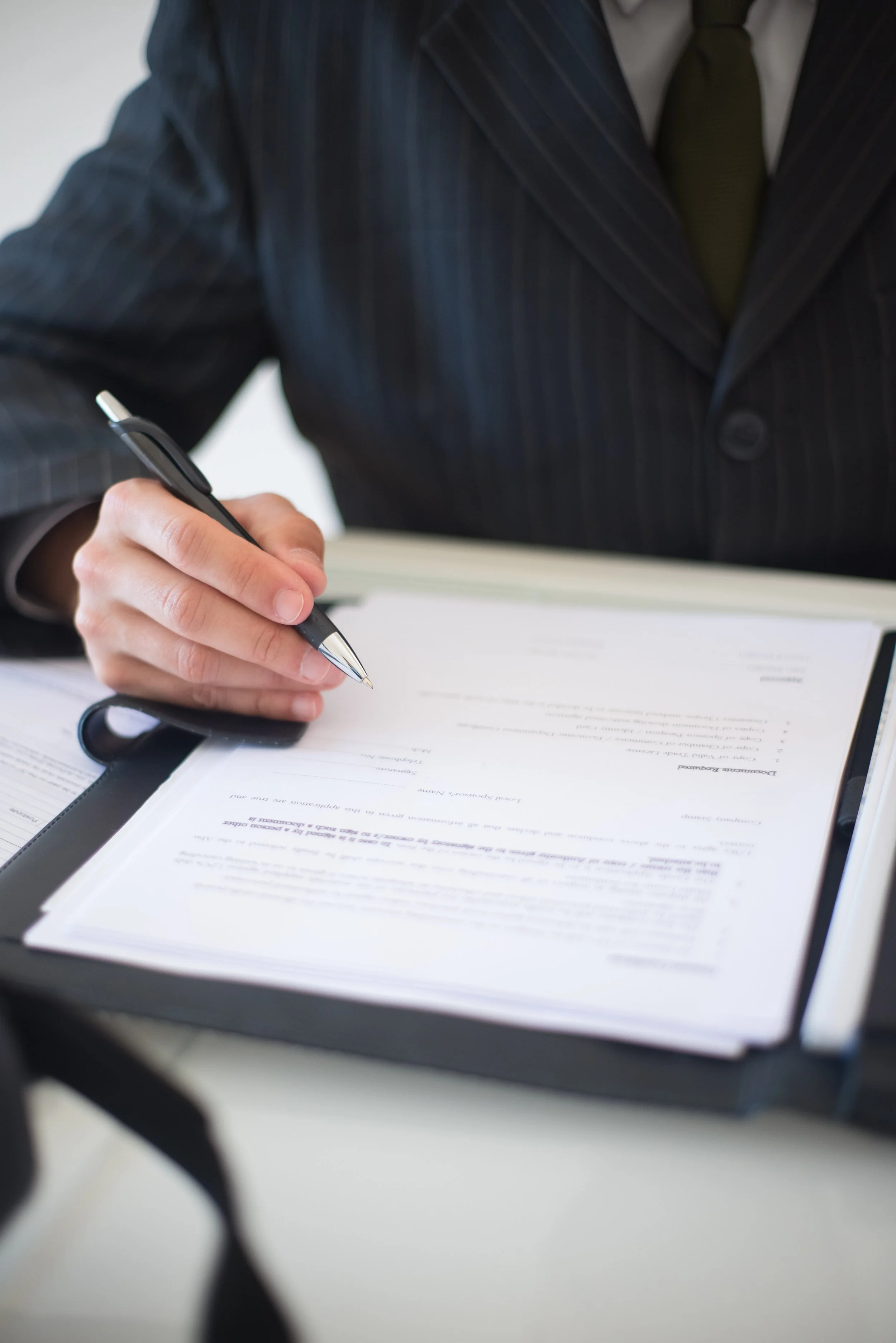 Person in a dark pinstripe suit and green tie signing documents with a black pen on a clipboard.