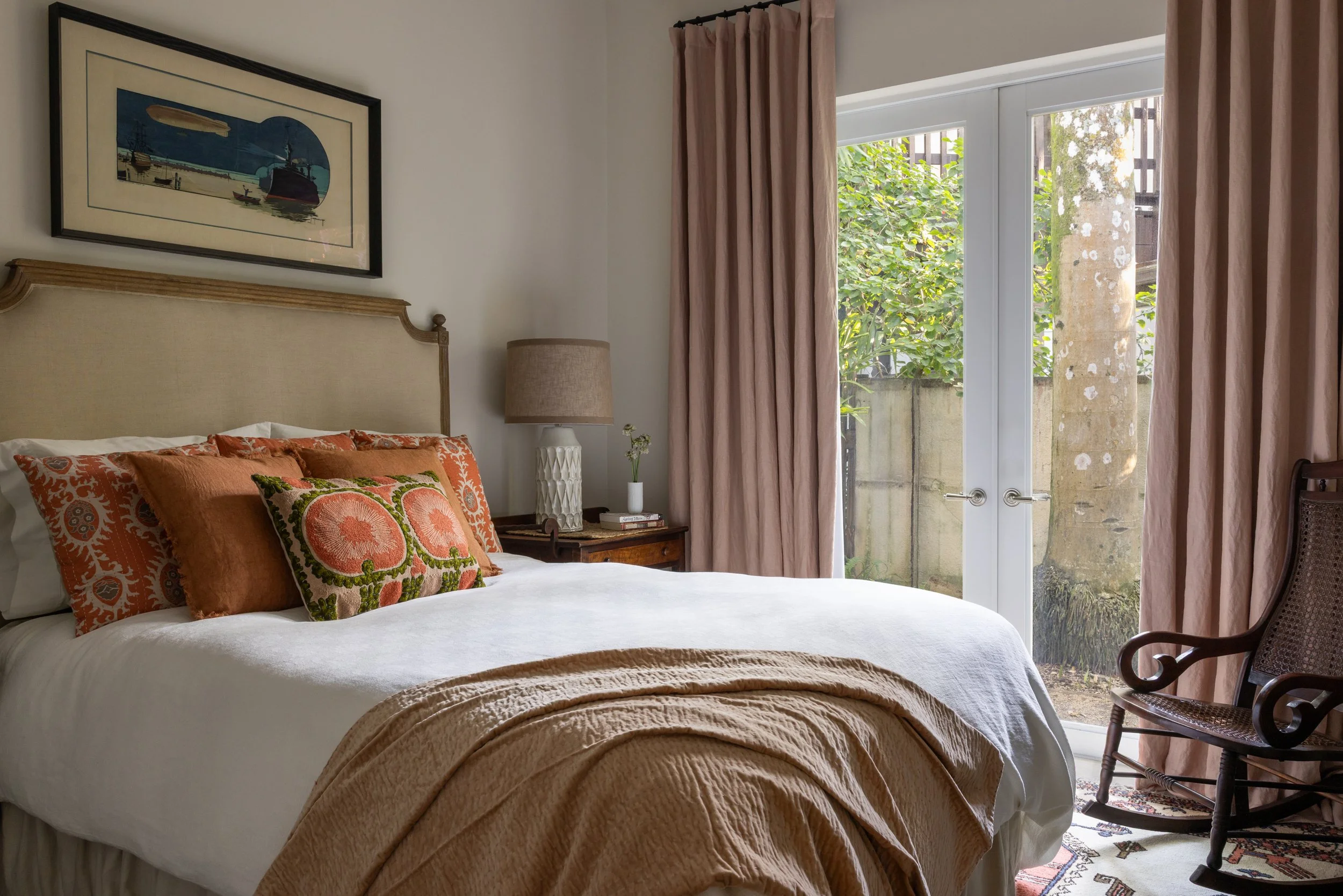 Charming guest bedroom with queen bed, paisley peach pillows, white duvet, muted orange coverlet, muted pink linen curtains, white textural bedside lamp with vintage wooden nightstand in Imparfait-designed space Crawford