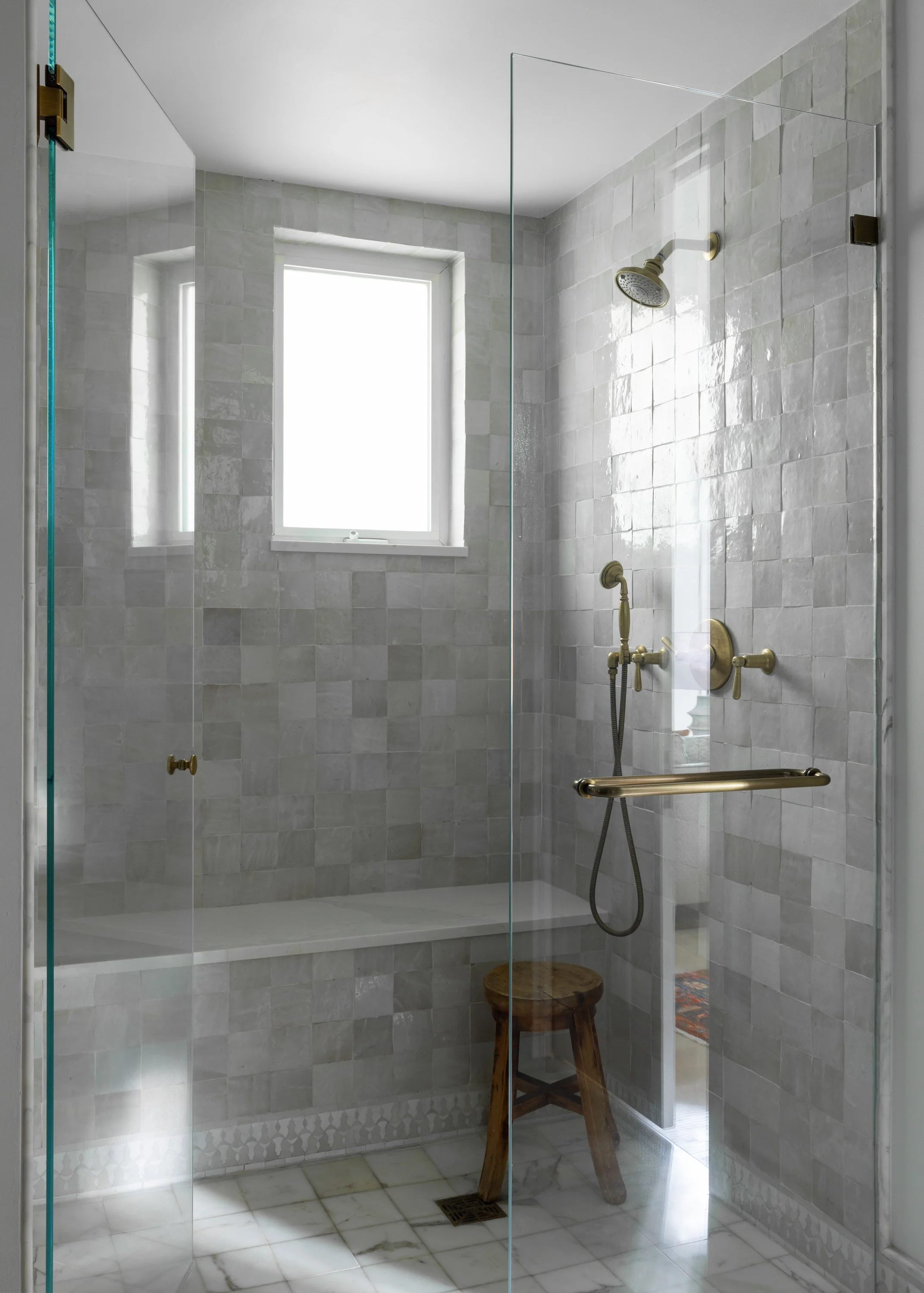 A sleek walk-in shower in the primary bathroom with a grey zellige tile bench with Moroccan tile border, white marble tiled floor, and brass faucet and knob pulls in Imparfait-designed space Crawford