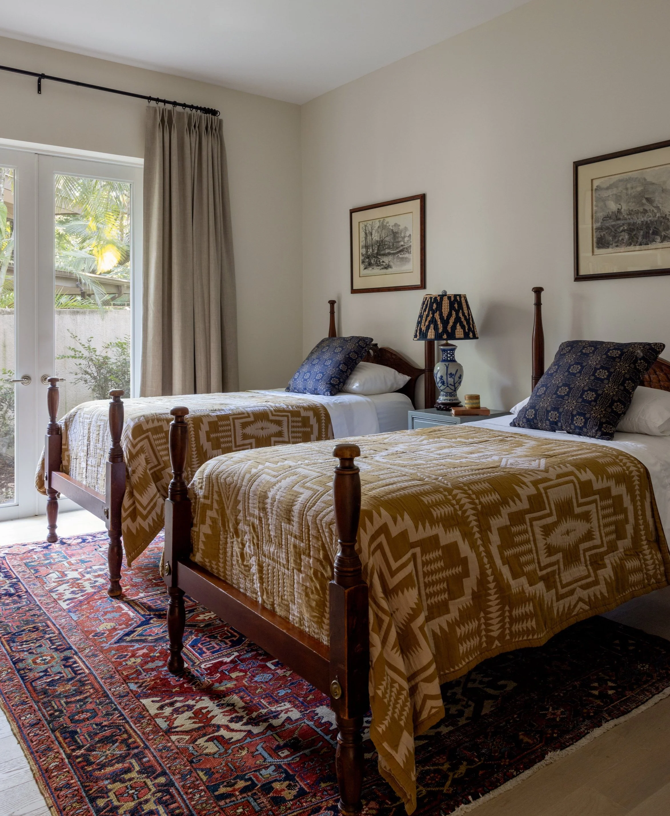 A cozy guest bedroom with twin poster beds, a red Persian rug, muted orange and pink aztec patterned coverlet, and blue and orange patterned pillows inside Imparfait-designed space Crawford