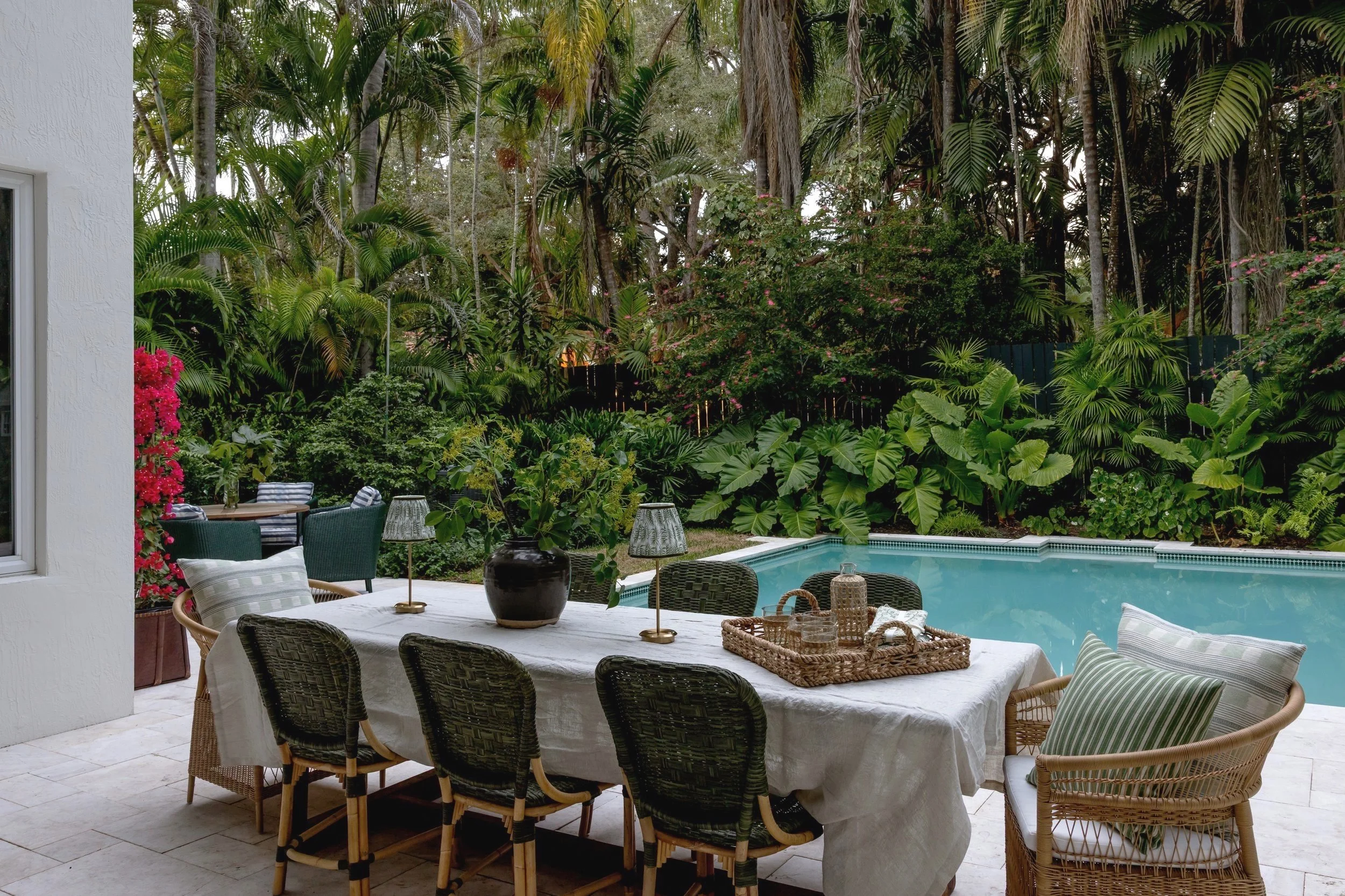 Outdoor patio with a dining table, surrounded by wicker chairs with cushions, next to a pool and lush green tropical plants and trees in the background.