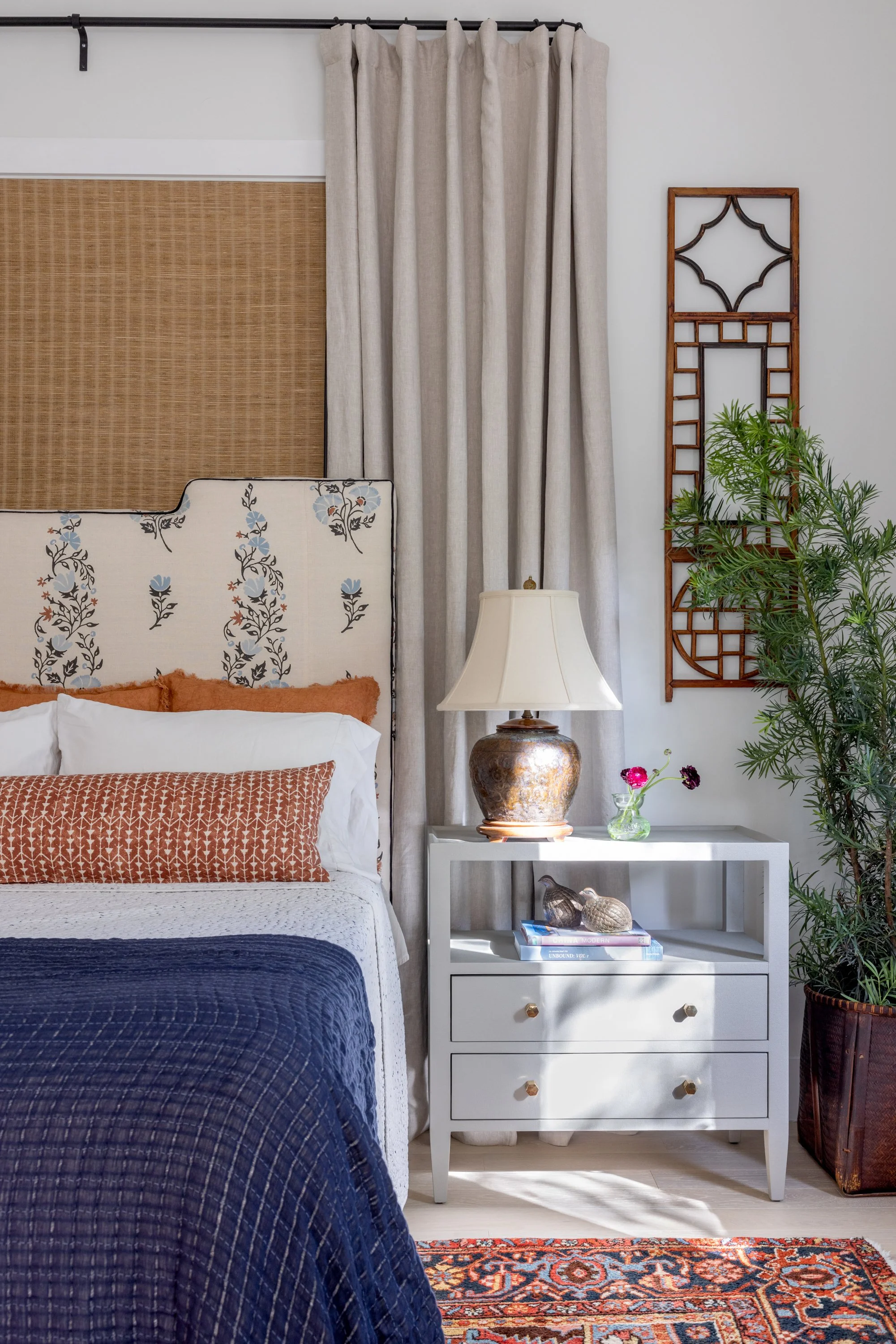 A vignette of the primary bedroom with a floral fabric headboard, layers of peachy orange pillows, a dark blue coverlet, a brass textural lamp, and light blue bedside table in Imparfait-designed space Crawford