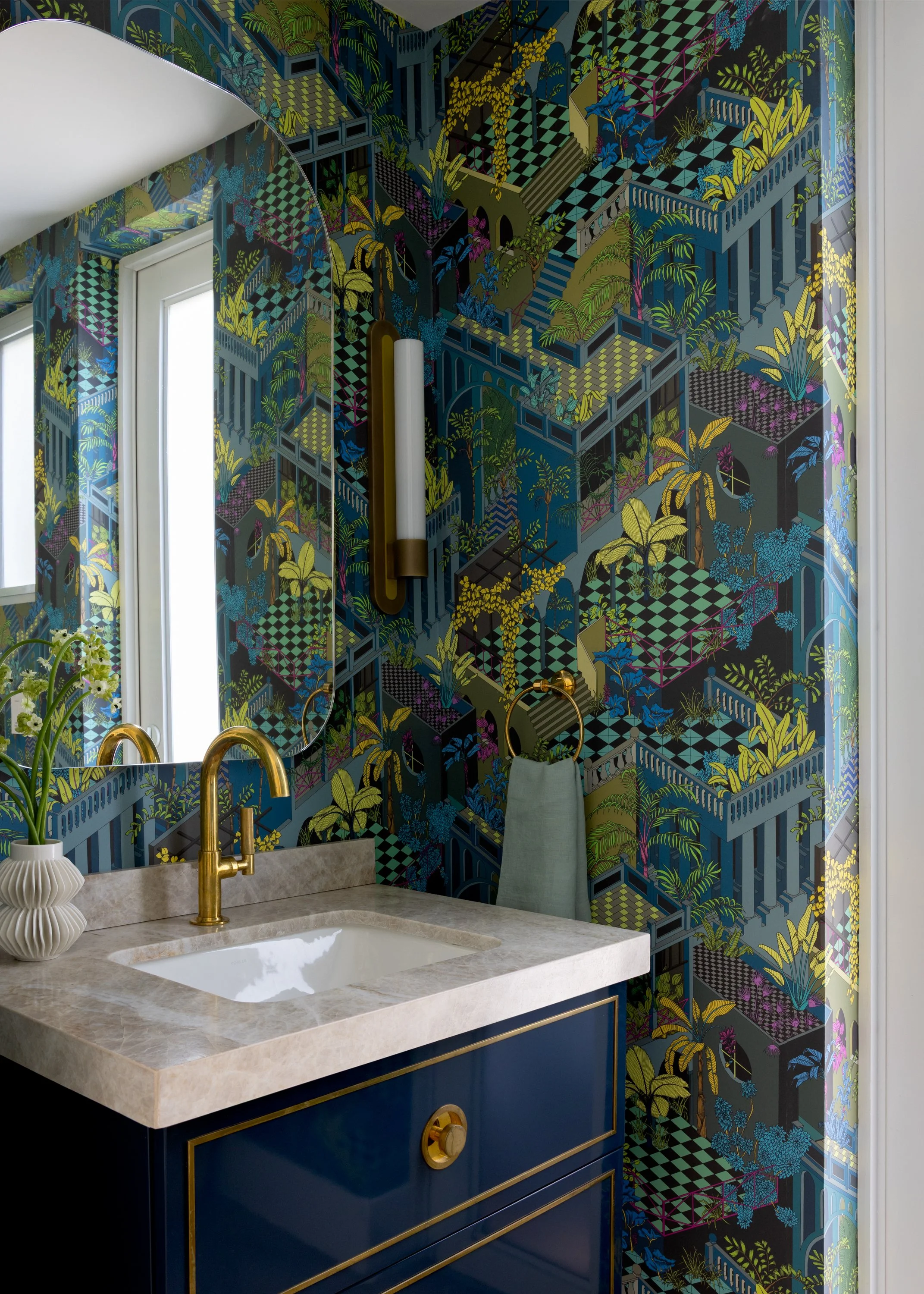 A vibrant powder room with a jungle-themed green and blue wallpaper, brass pill sconces, a high gloss dark blue vanity with a cream colored marble countertop, and brass lacquered faucet in Imparfait-designed space Crawford