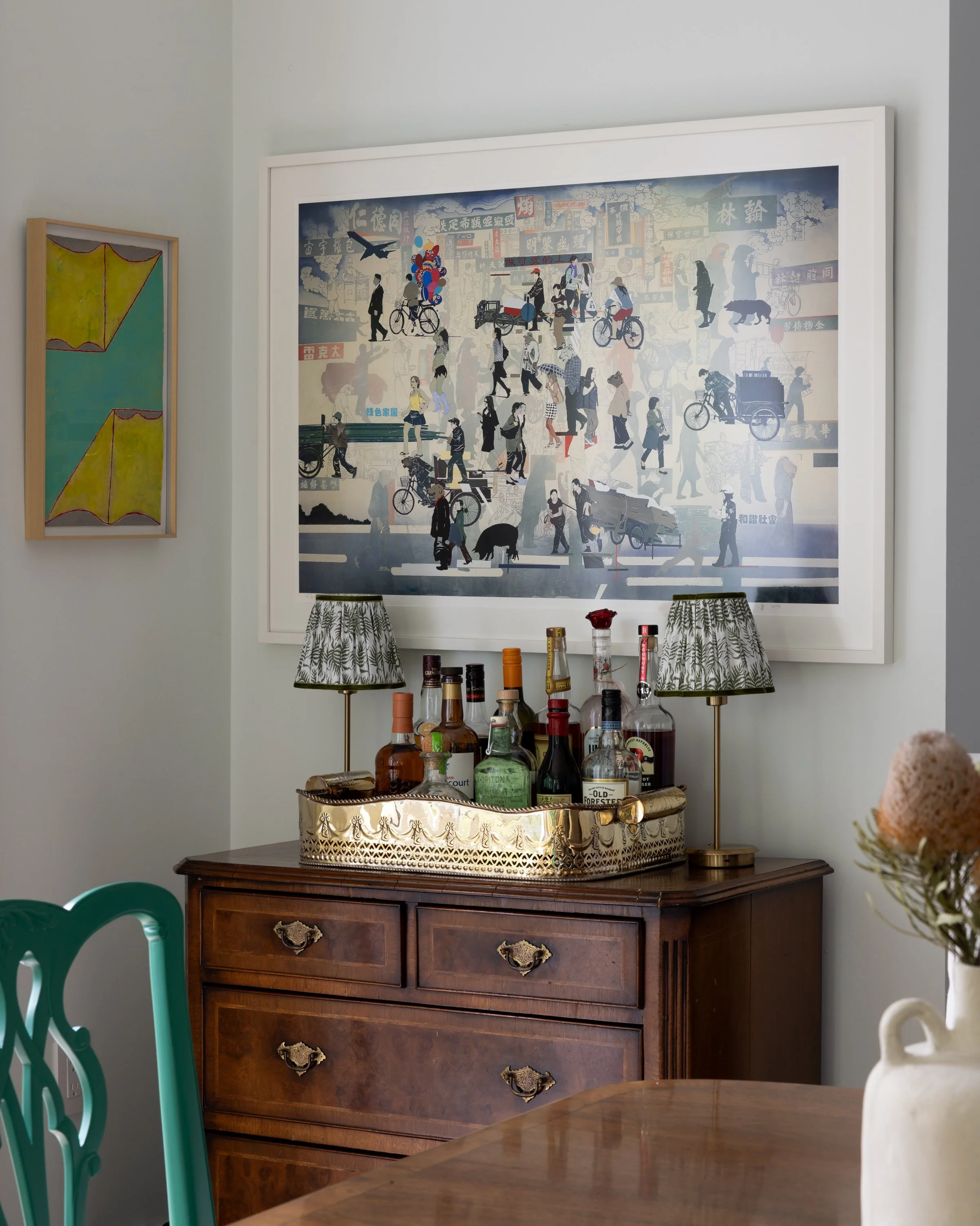 Bar nook with beautiful art, a brass tray with bar items, Ming cabinet, and two green and white paisley portable lamps inside Imparfait Design Studio-designed Crawford