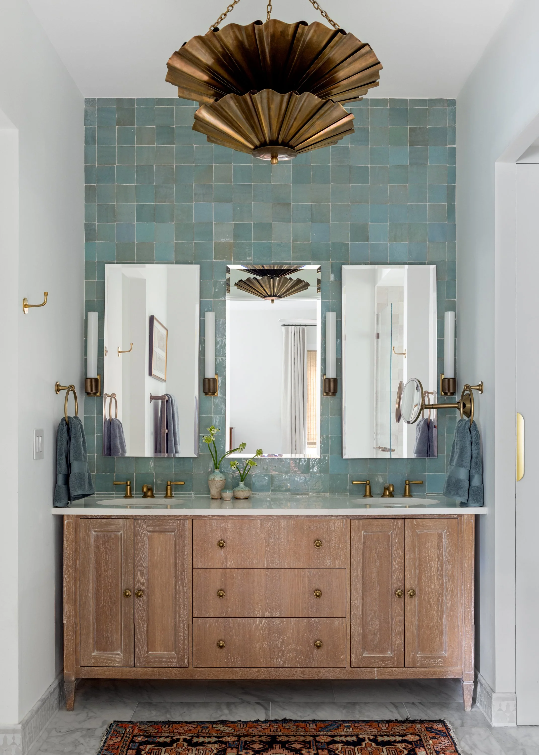 A gorgeous master bath with a brass flowering chandelier, brass and lucite sconces, ocean blue Zellige tile, brass unlacquered towel bars and faucets, and a distressed white oak double vanity in Imparfait-designed space Crawford