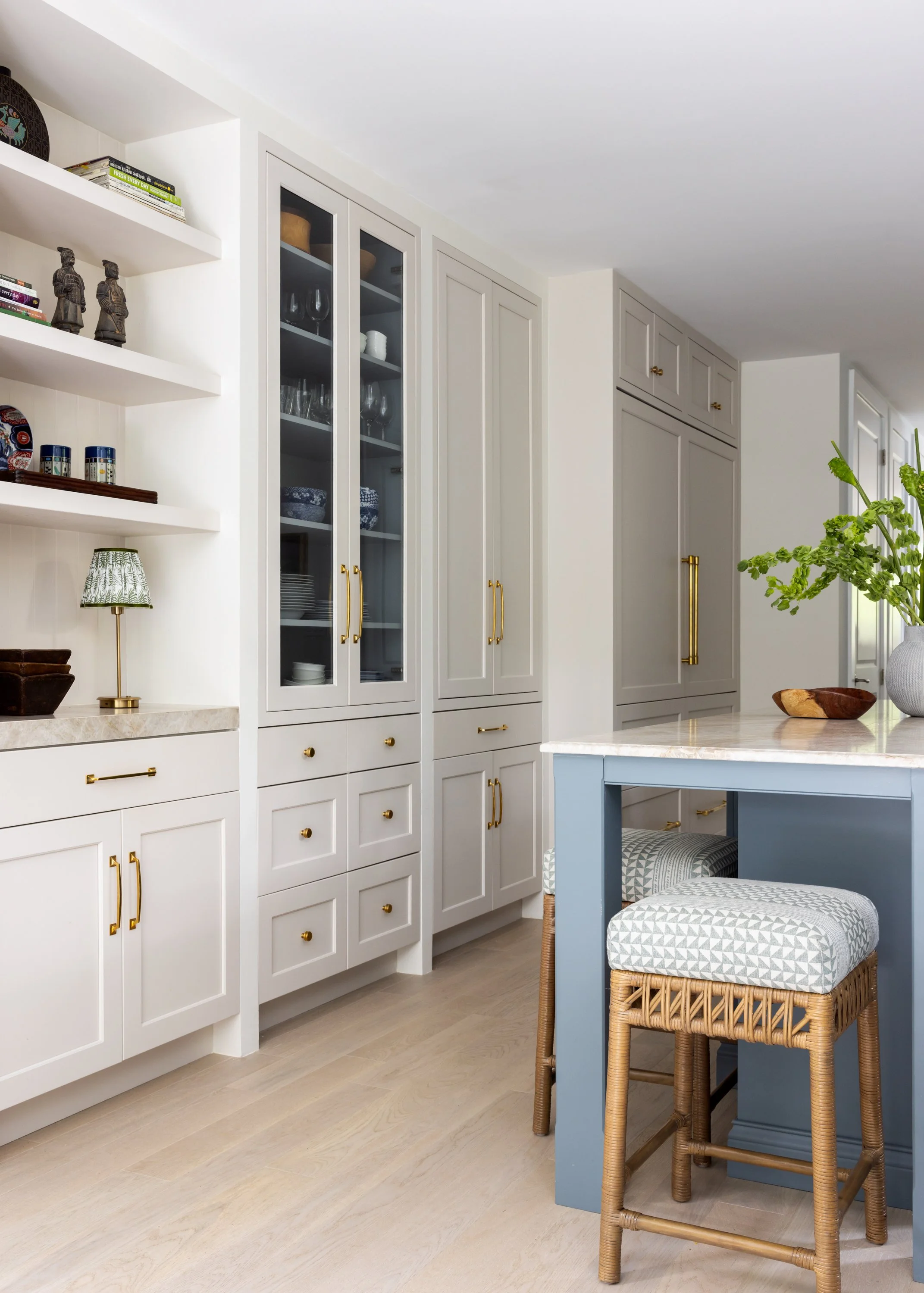 Serene view of the kitchen with low to ceiling white cabinetry, an art niche, a blue center kitchen island with beige marble countertops, and rattan counter stools with patterned light blue cushions inside Imparfait-designed space Crawford