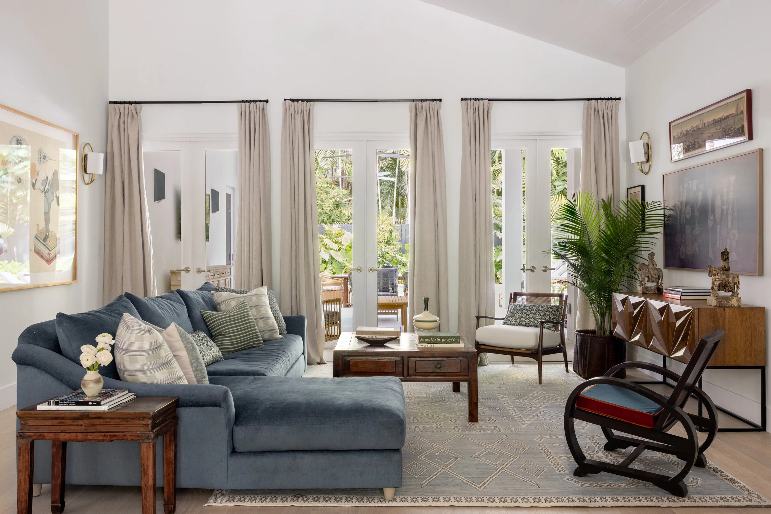 Main living room with velvet deep blue sofa, Ming coffee table and side tables, a rattan curved lounge chair, and black bentwood rocking chair with a blue and red cushion in Imparfait-designed space Crawford